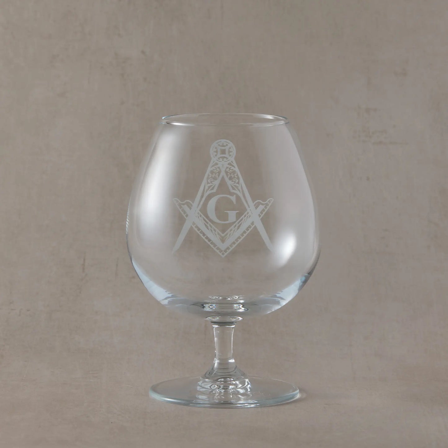 Clear brandy glass engraved with the Masonic square and compass with G symbol, photographed on a soft neutral beige background
