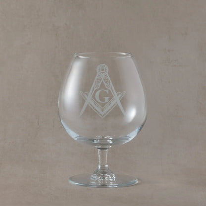 Clear brandy glass engraved with the Masonic square and compass with G symbol, photographed on a soft neutral beige background