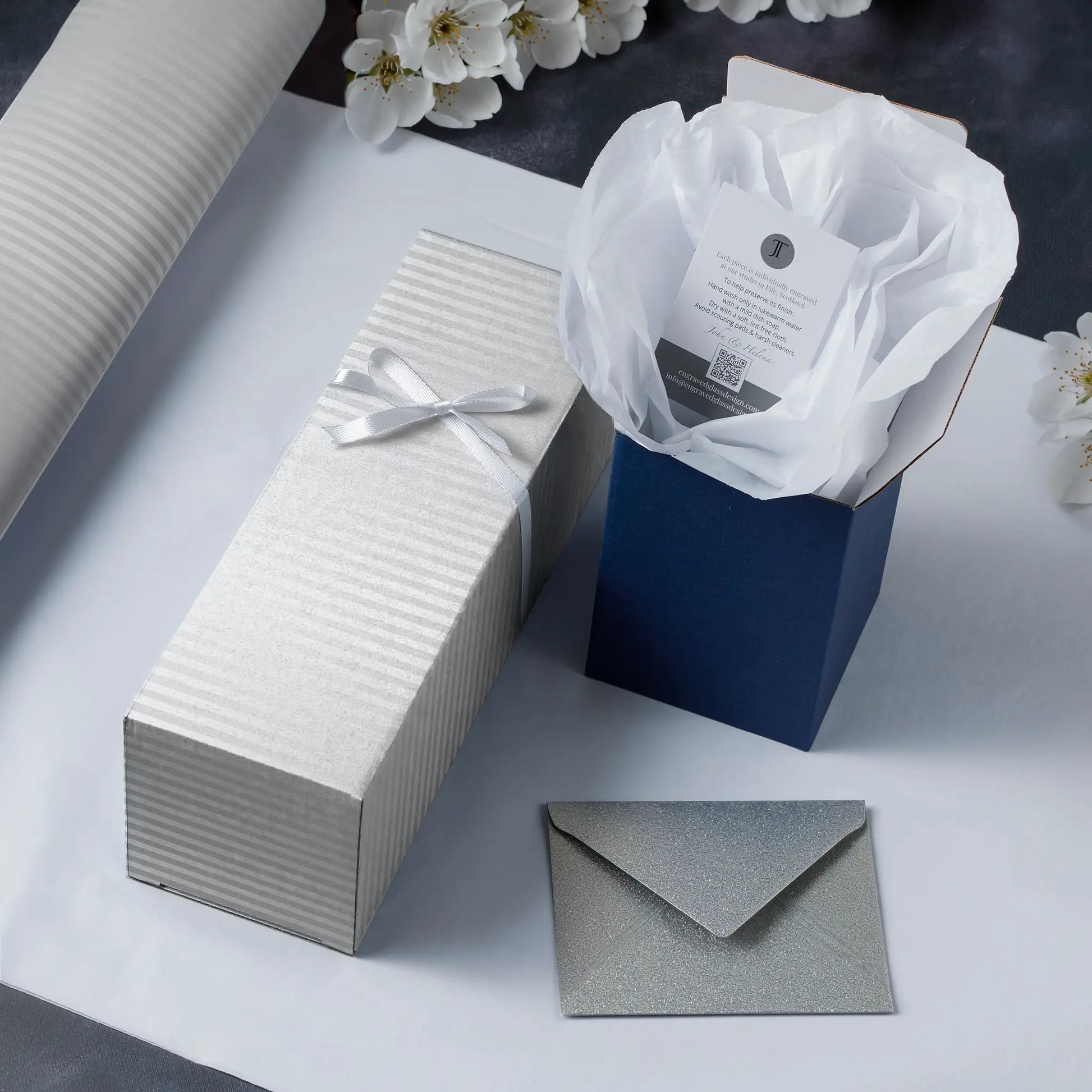 Gift box, tissue paper in a blue box, and silver envelope on a light surface with flowers in the background.