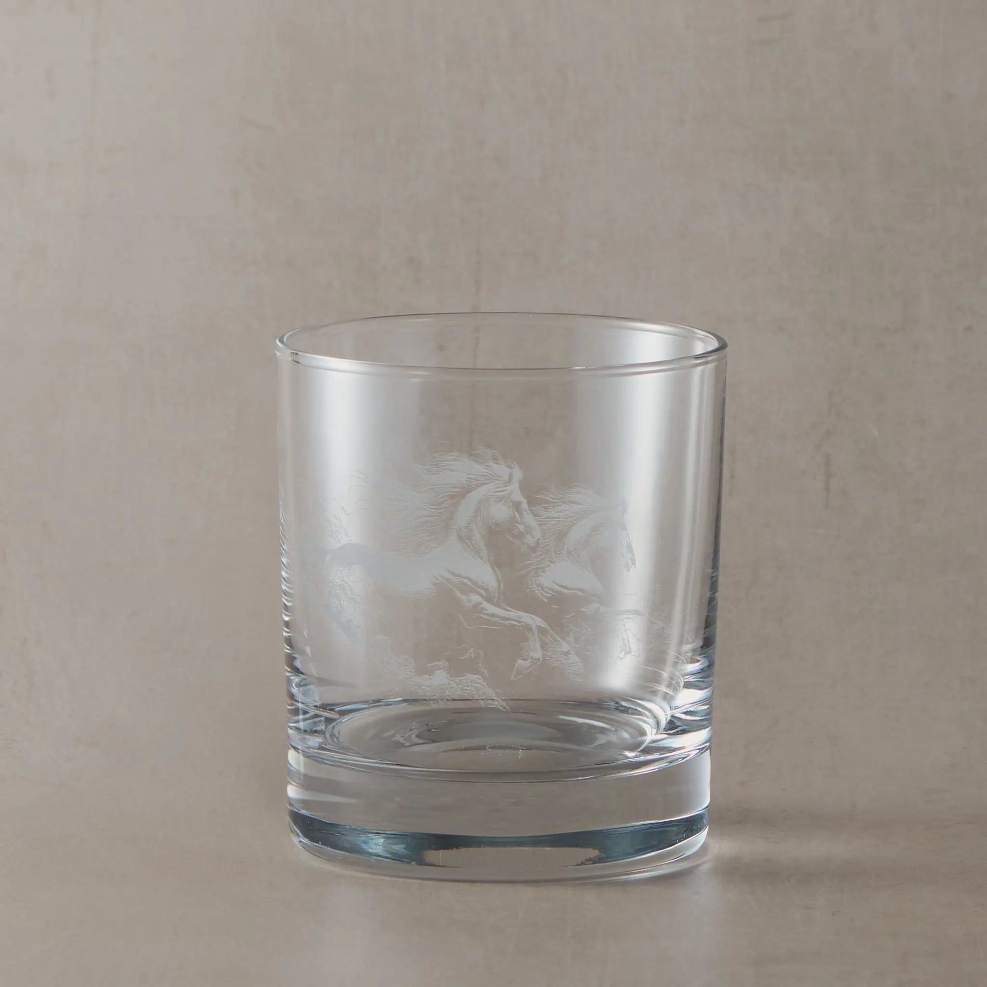 Clear whisky glass engraved with two galloping unicorns, photographed on a soft neutral beige background