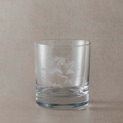 Clear whisky glass engraved with two galloping unicorns, photographed on a soft neutral beige background
