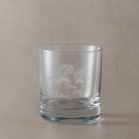 Clear whisky glass engraved with two galloping unicorns, photographed on a soft neutral beige background