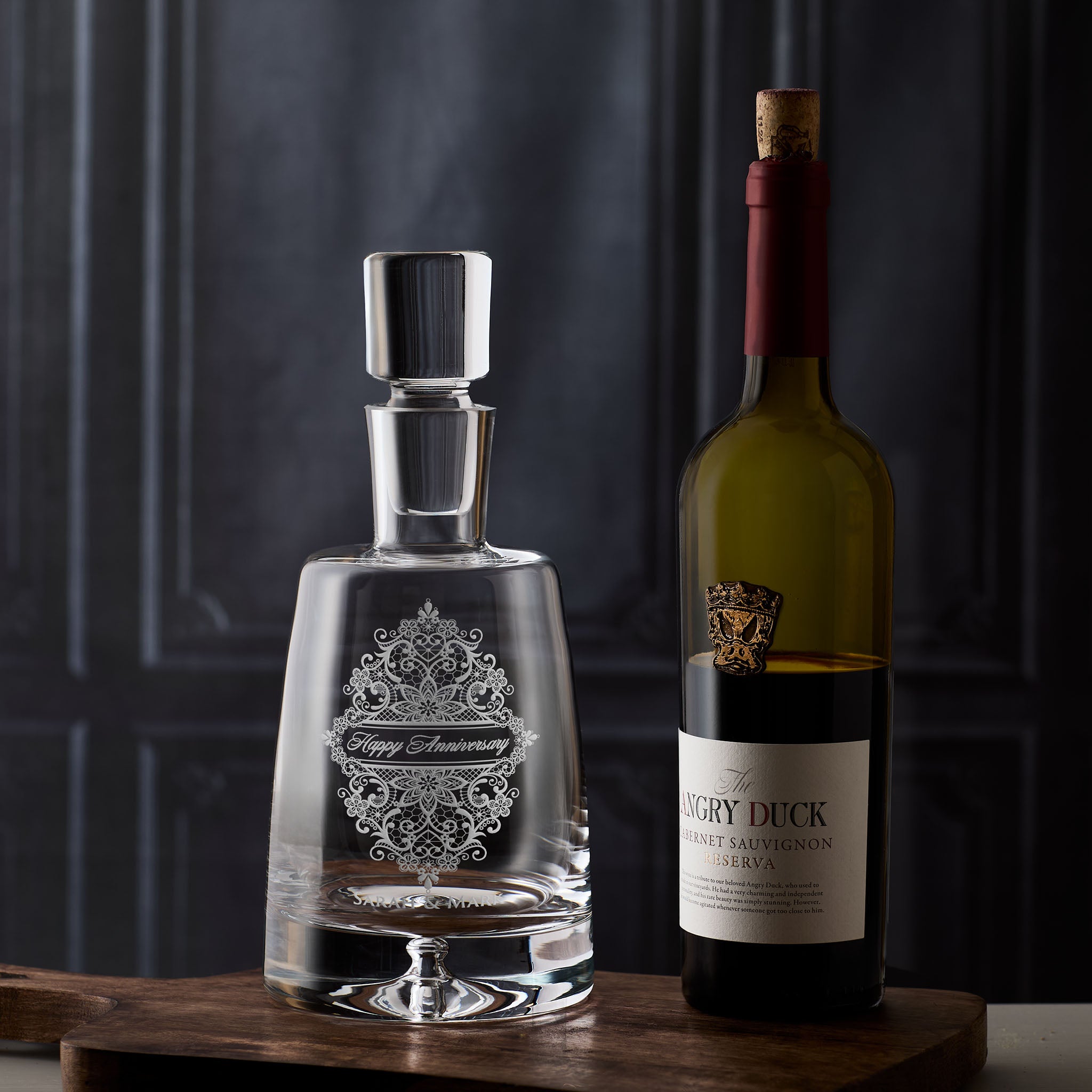 25th Wedding Anniversary decanter with decorative floral lacework engraving next to a wine bottle on a wooden surface.