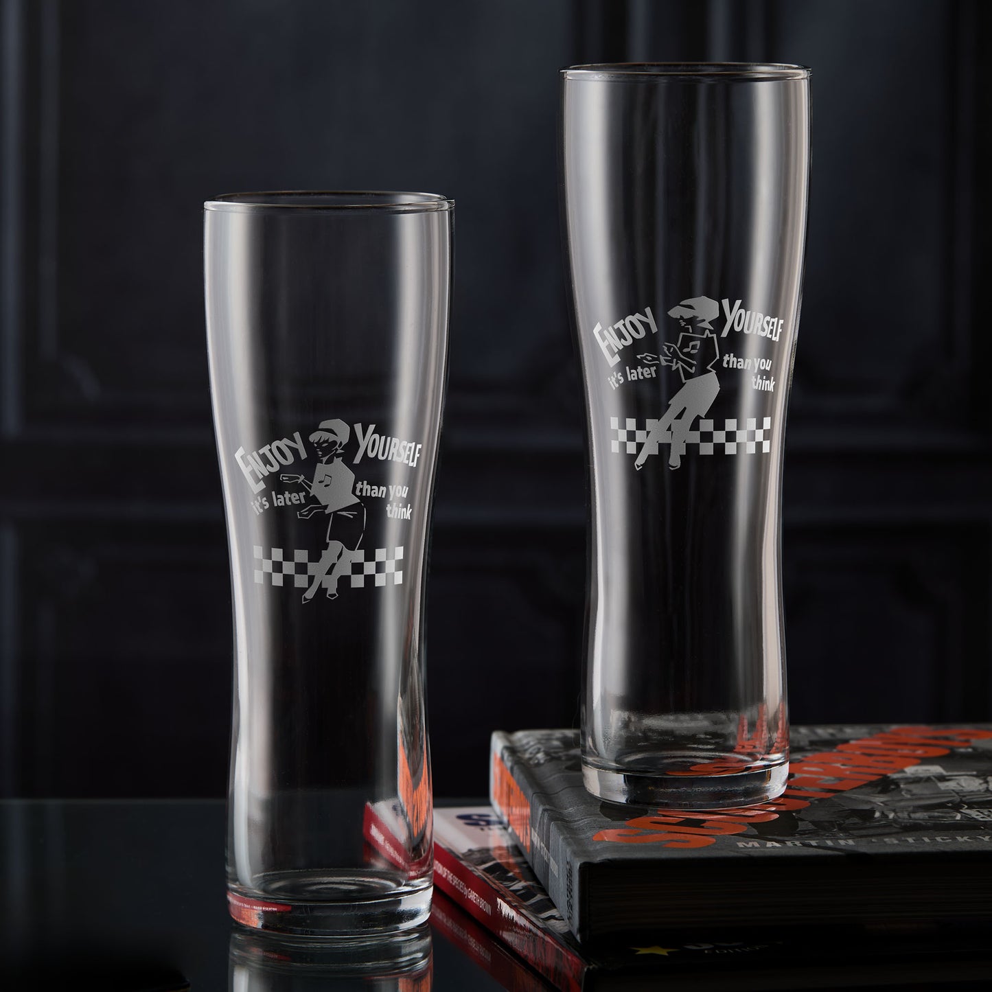 Two engraved Beat Girl pint glasses with “Enjoy Yourself” text and checkerboard design.