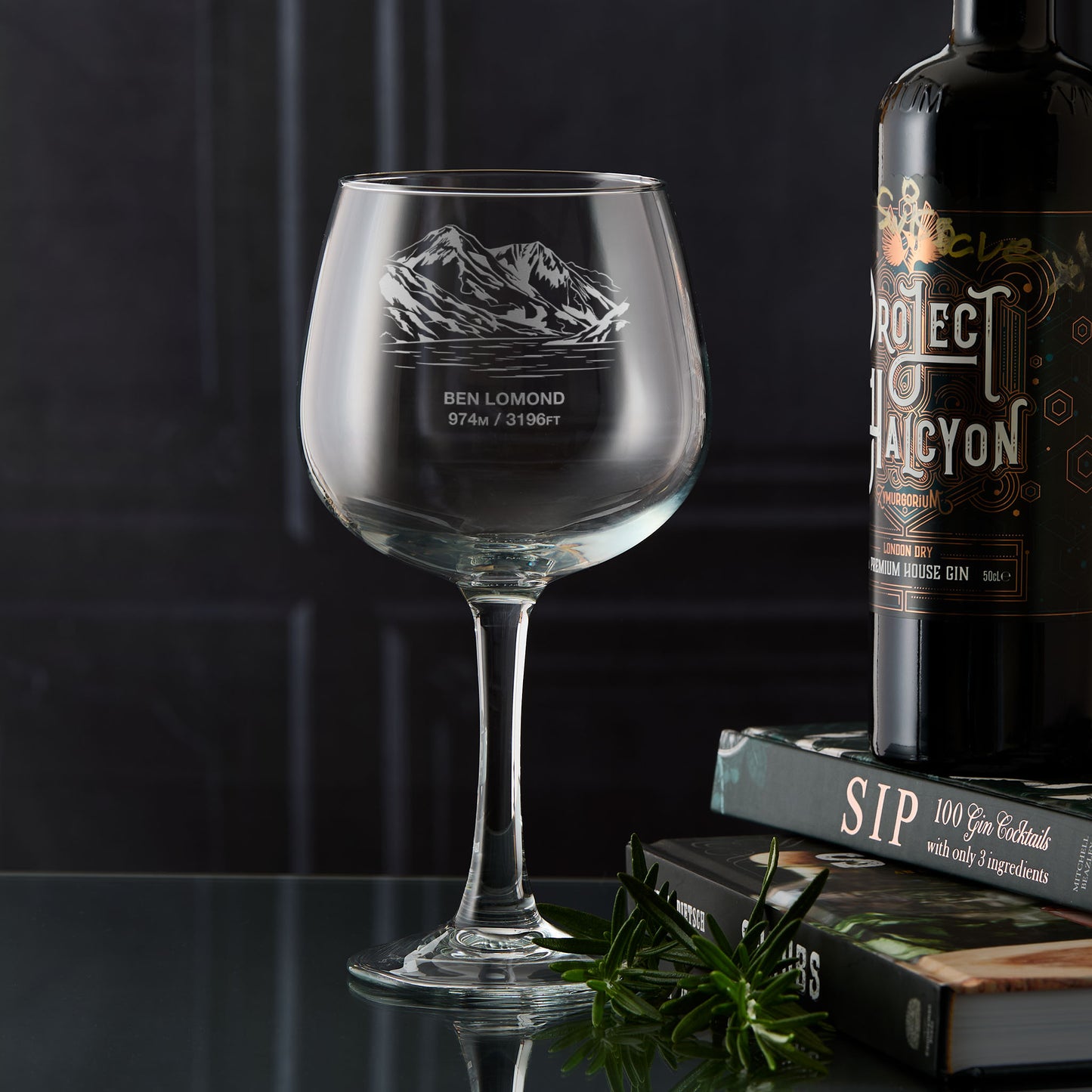 Engraved gin glass with the mountain Ben Lomond next to a bottle of gin on books against a dark background