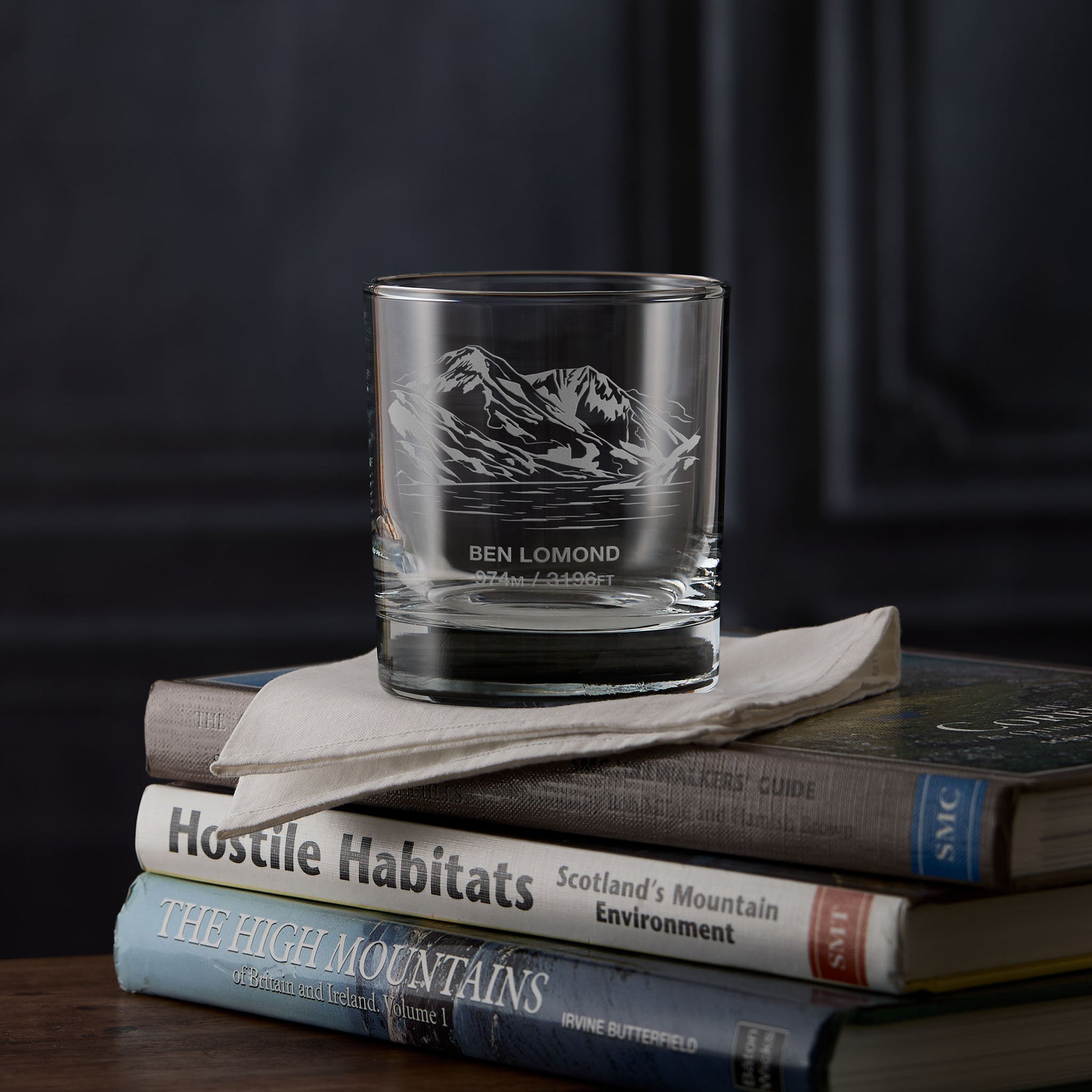 Whisky glass engraved with the mountain Ben Lomond on a stack of books against a dark background