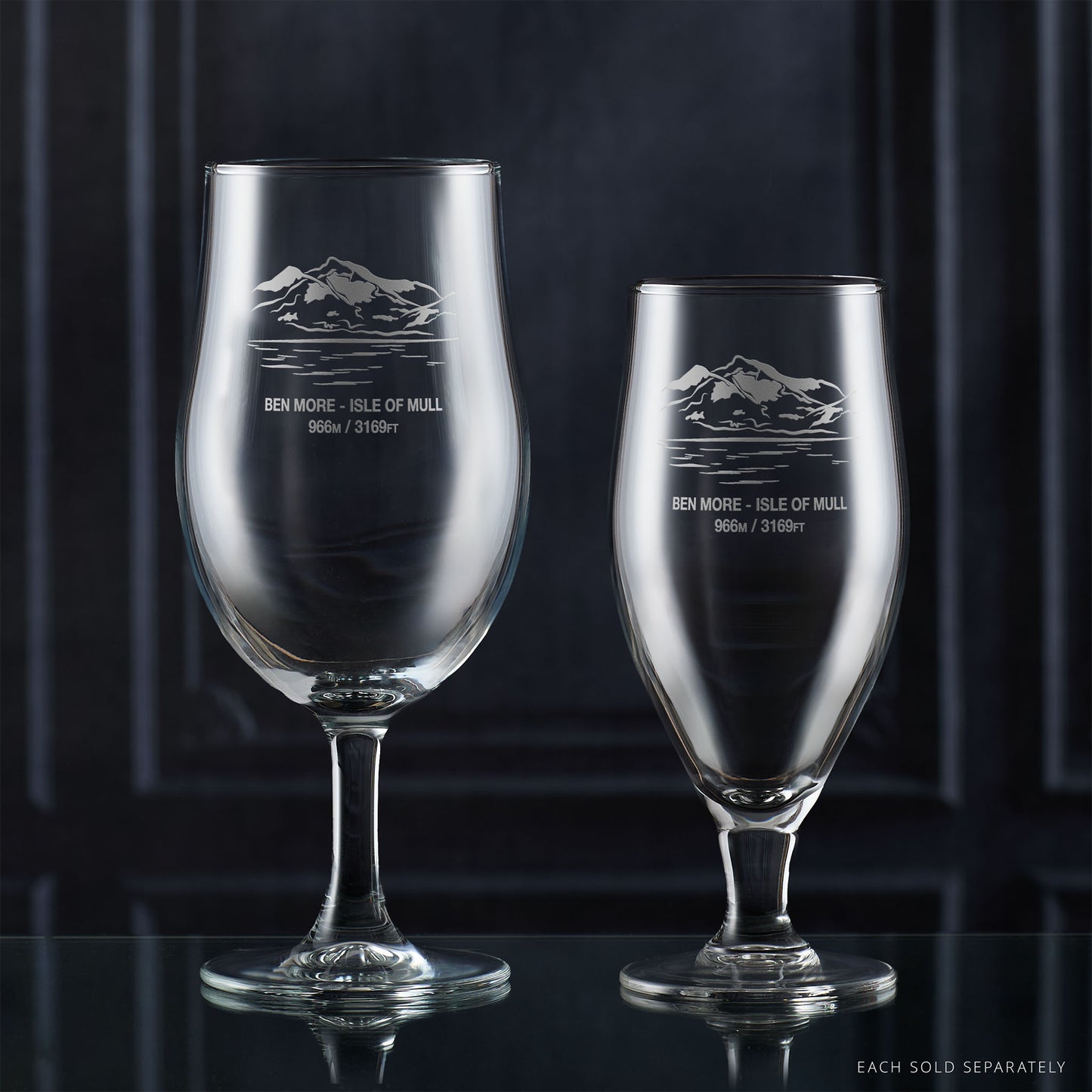 Two stemmed beer glasses with engraved mountain design on a dark background