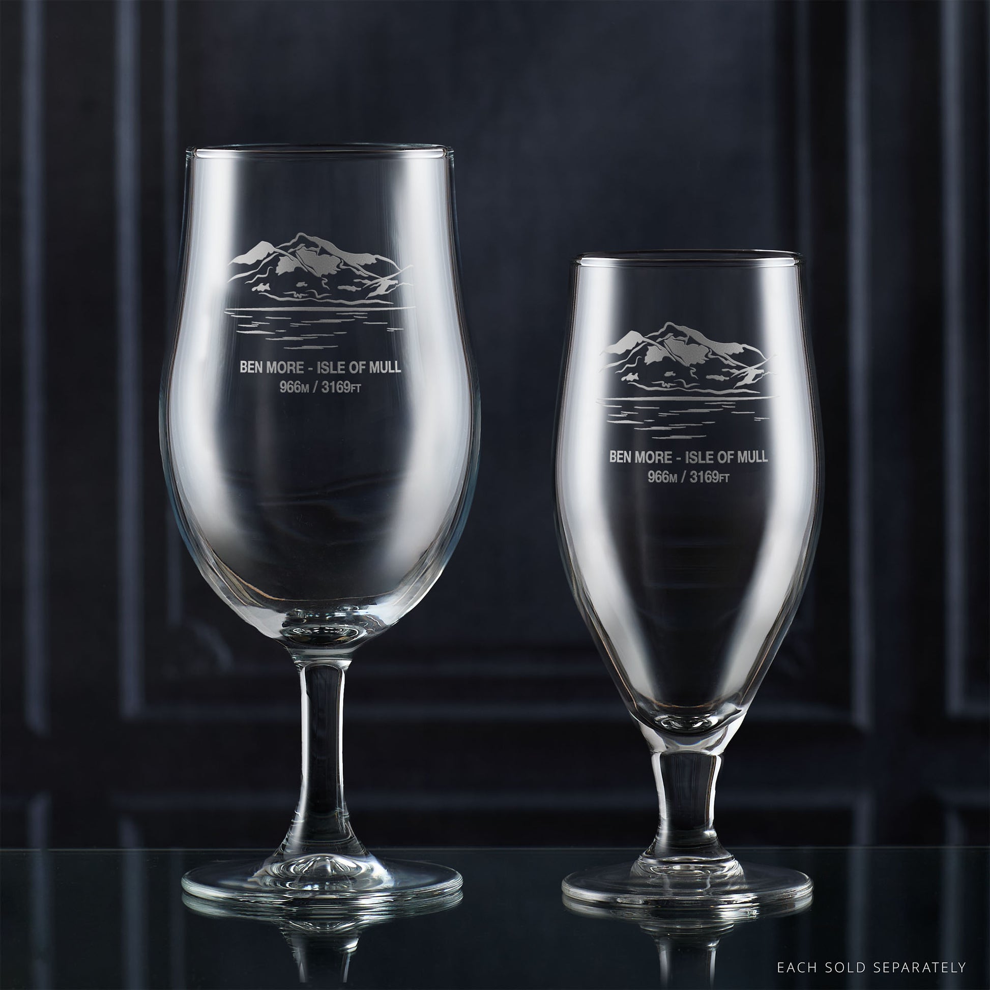 Two stemmed beer glasses with engraved mountain design on a dark background