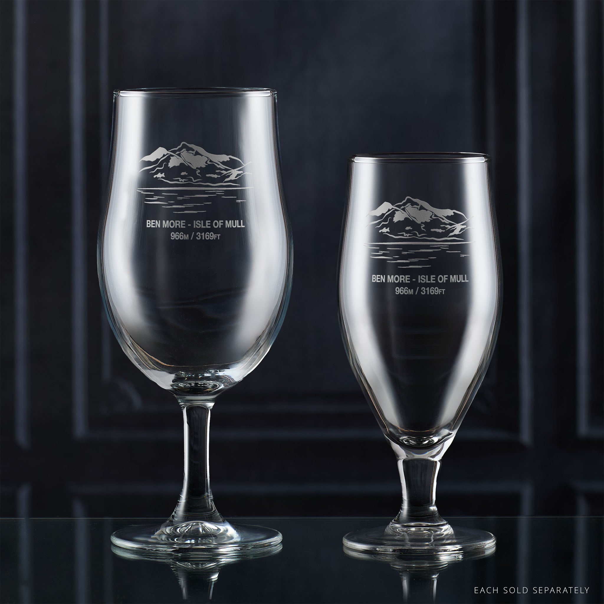 Two stemmed beer glasses with engraved mountain design on a dark background