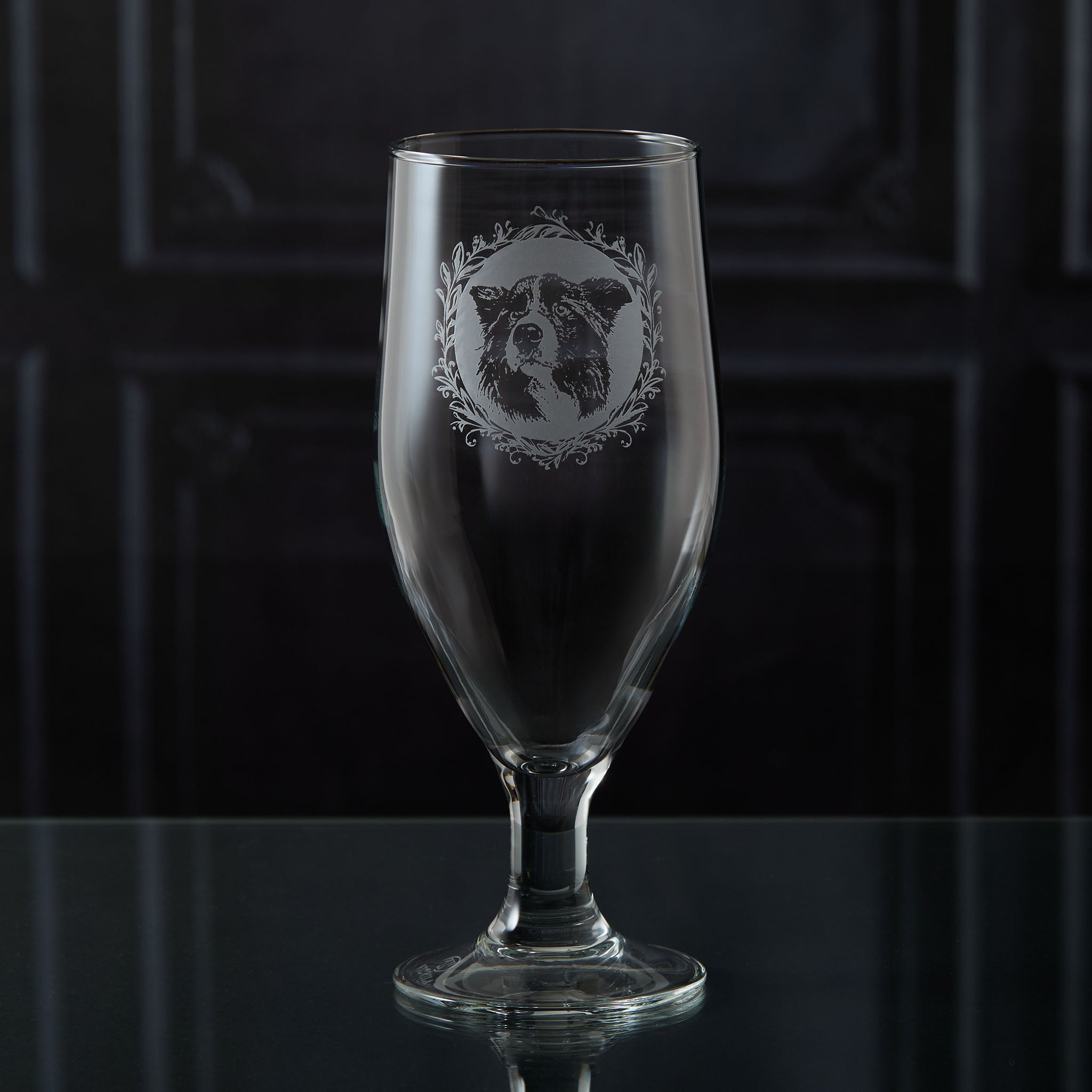 Stemmed beer glass with engraved design of a Border Collie's face on a dark background