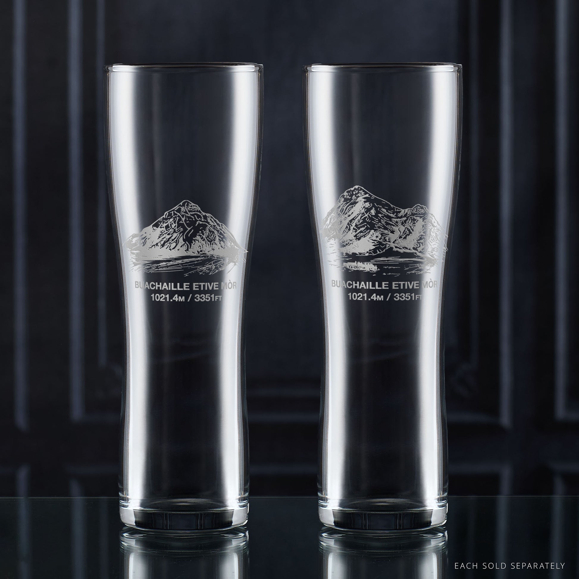Two clear pint glasses with mountain etching on a dark background