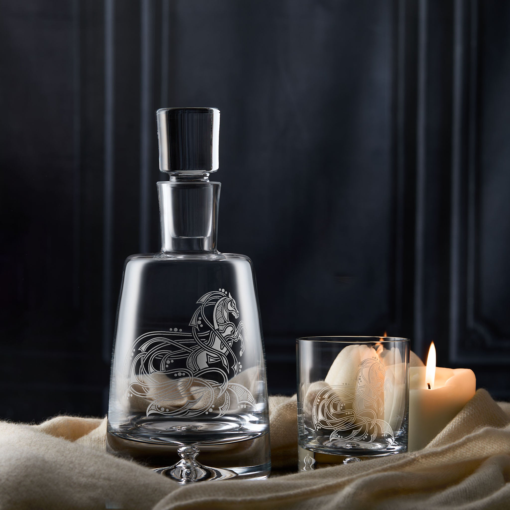 Glass decanter with engraved horse design next to a whisky glass with a candle on a dark background