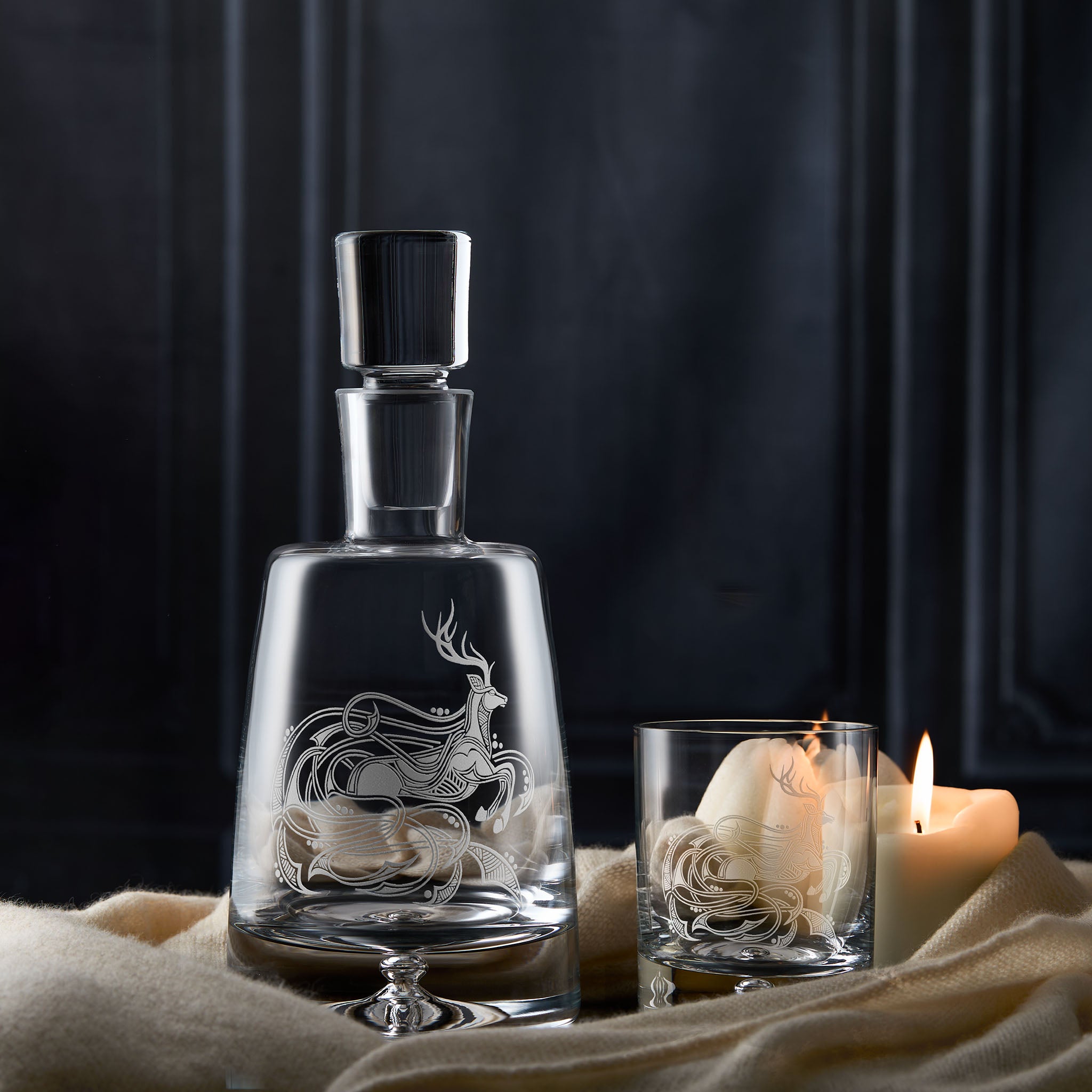 Highland stag decanter with engraved deer design next to a whisky glass with a candle on a dark background