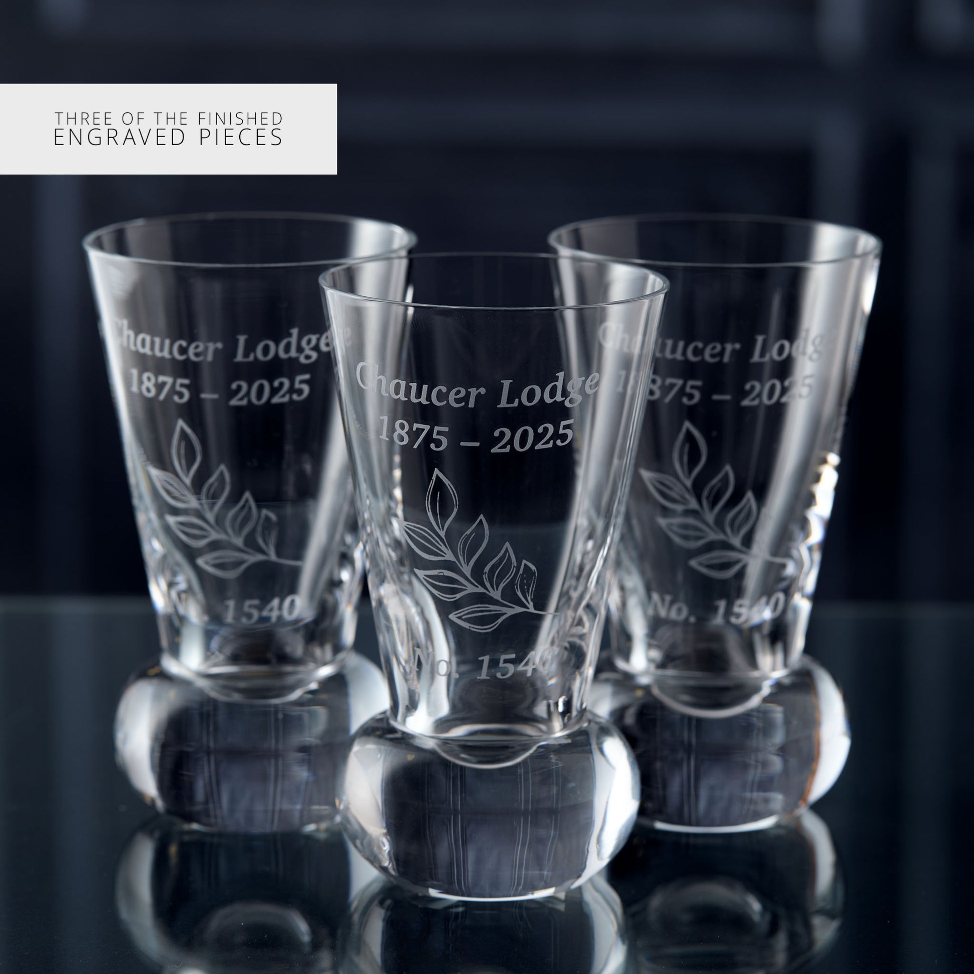 Three engraved glass tumblers on a reflective surface with a dark background