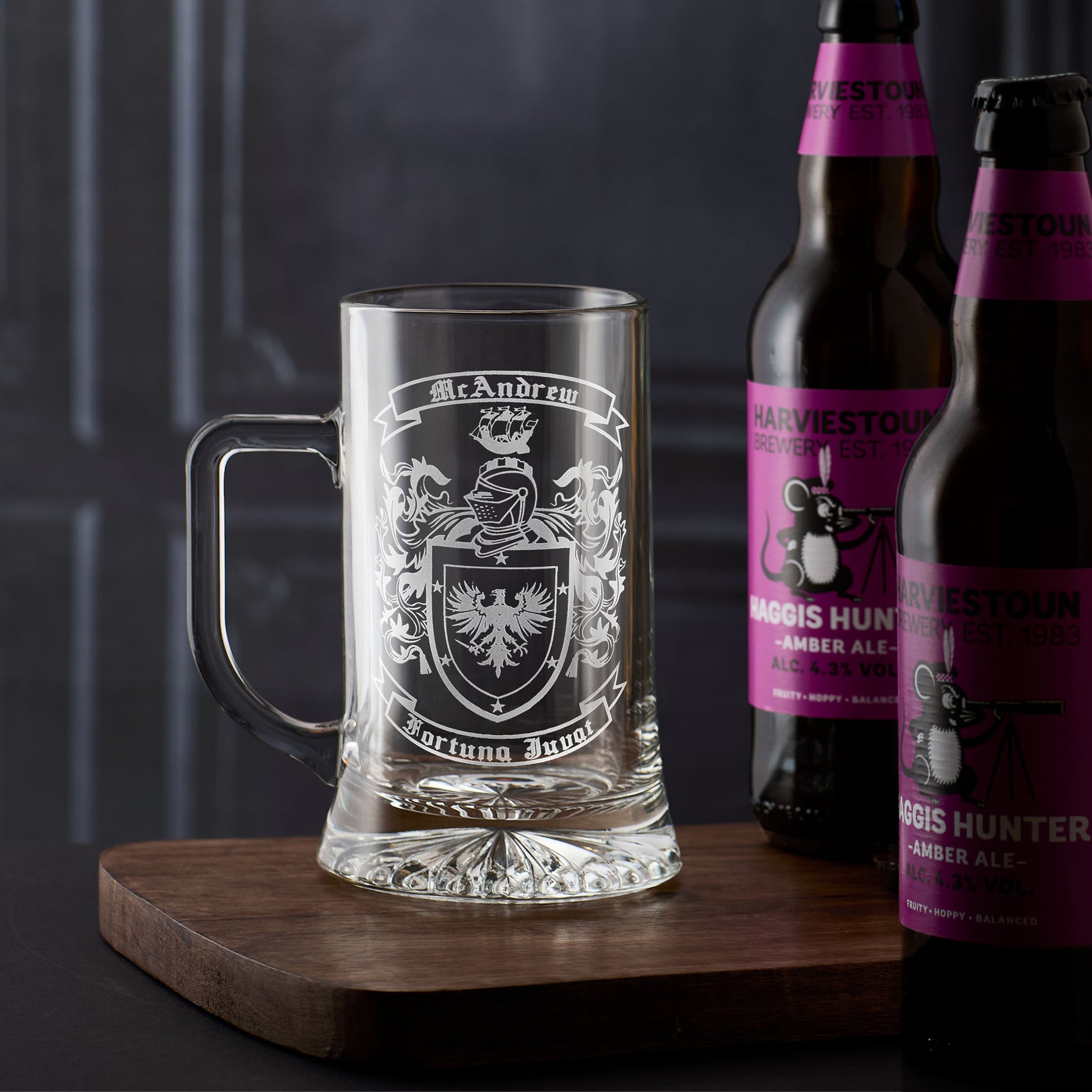 Glass tankard with engraved coat of arms heraldry design and two bottles of beer on a wooden board.