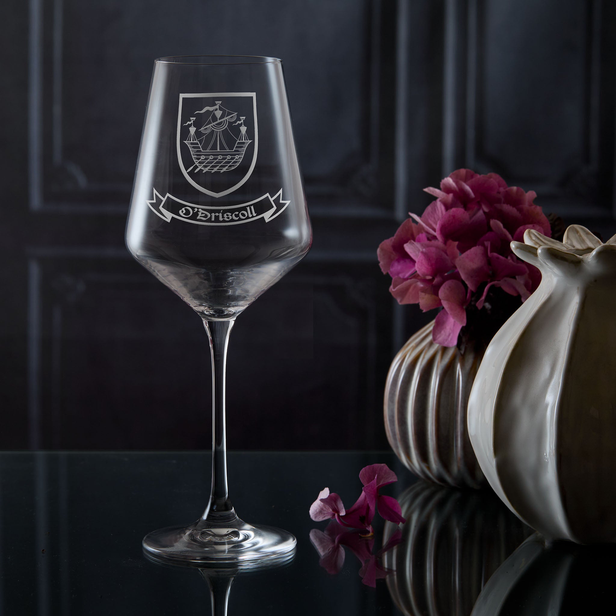 Wine glass with engraved coat of arms design on a dark surface with flowers and a vase.