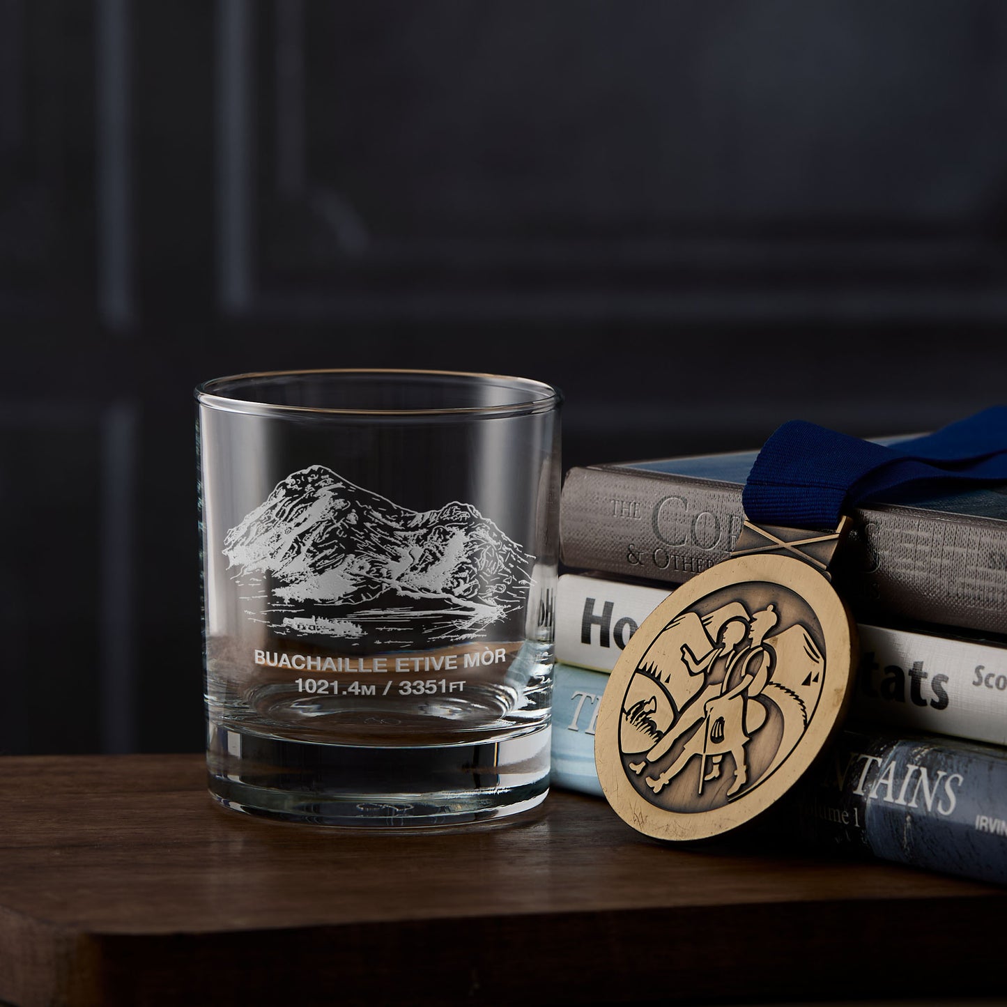 whisky glass with Buachaille Etive Mor mountain engraving, books, and a hillwalking medal on a wooden surface