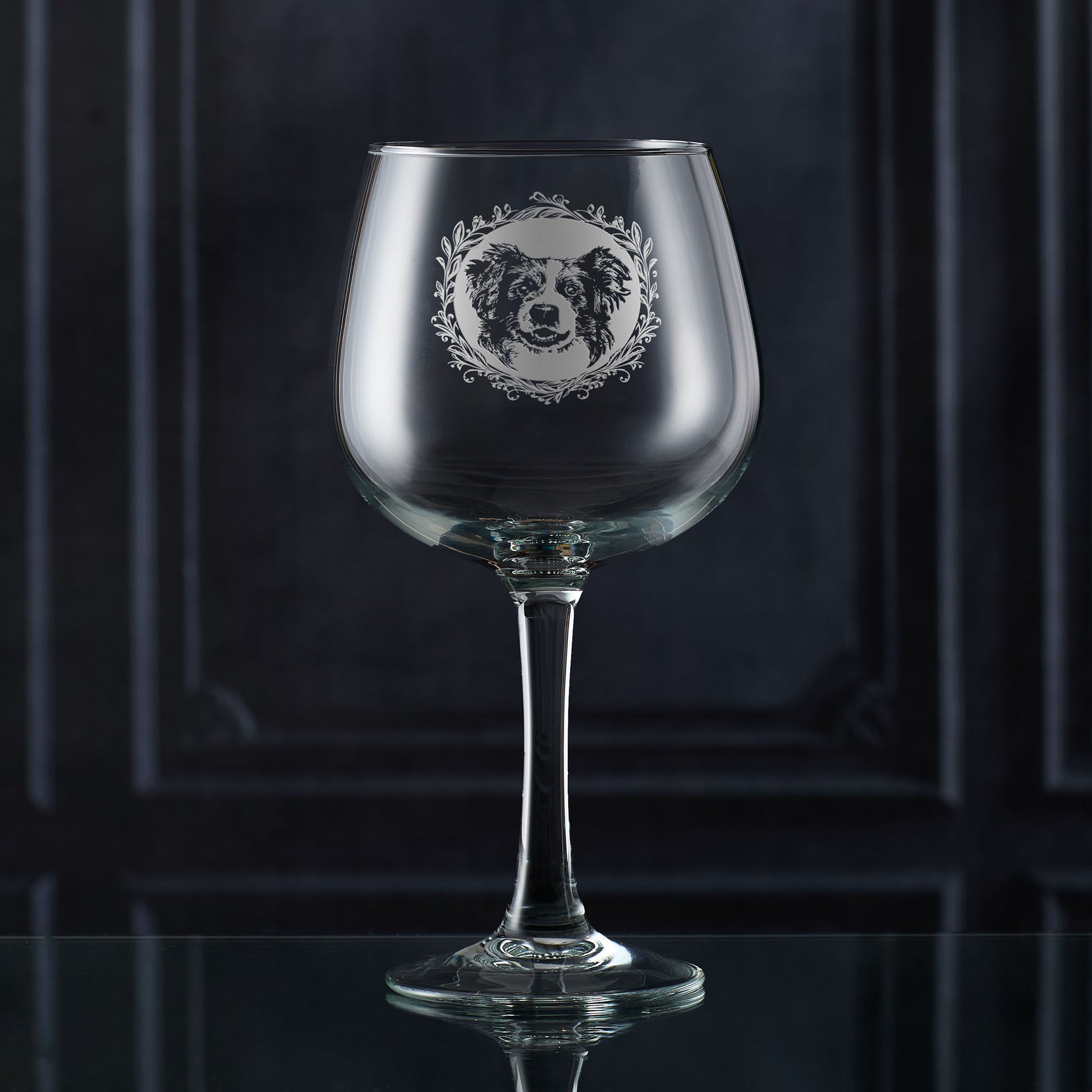 Hand-engraved Border Collie gin glass shown in front of a dark panelled wall, featuring a leafy garland and detailed Collie portrait design by Just The Ticket.