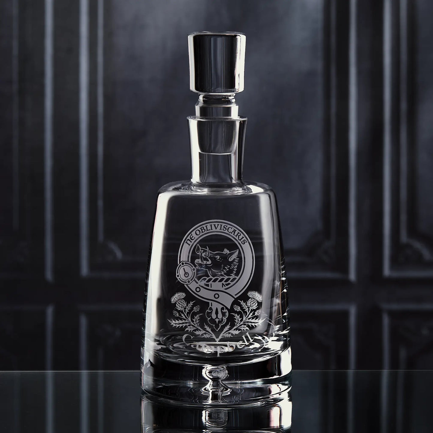 Handmade glass decanter engraved with personalised clan crest and thistle motif, shown on dark background to highlight shape and craftsmanship