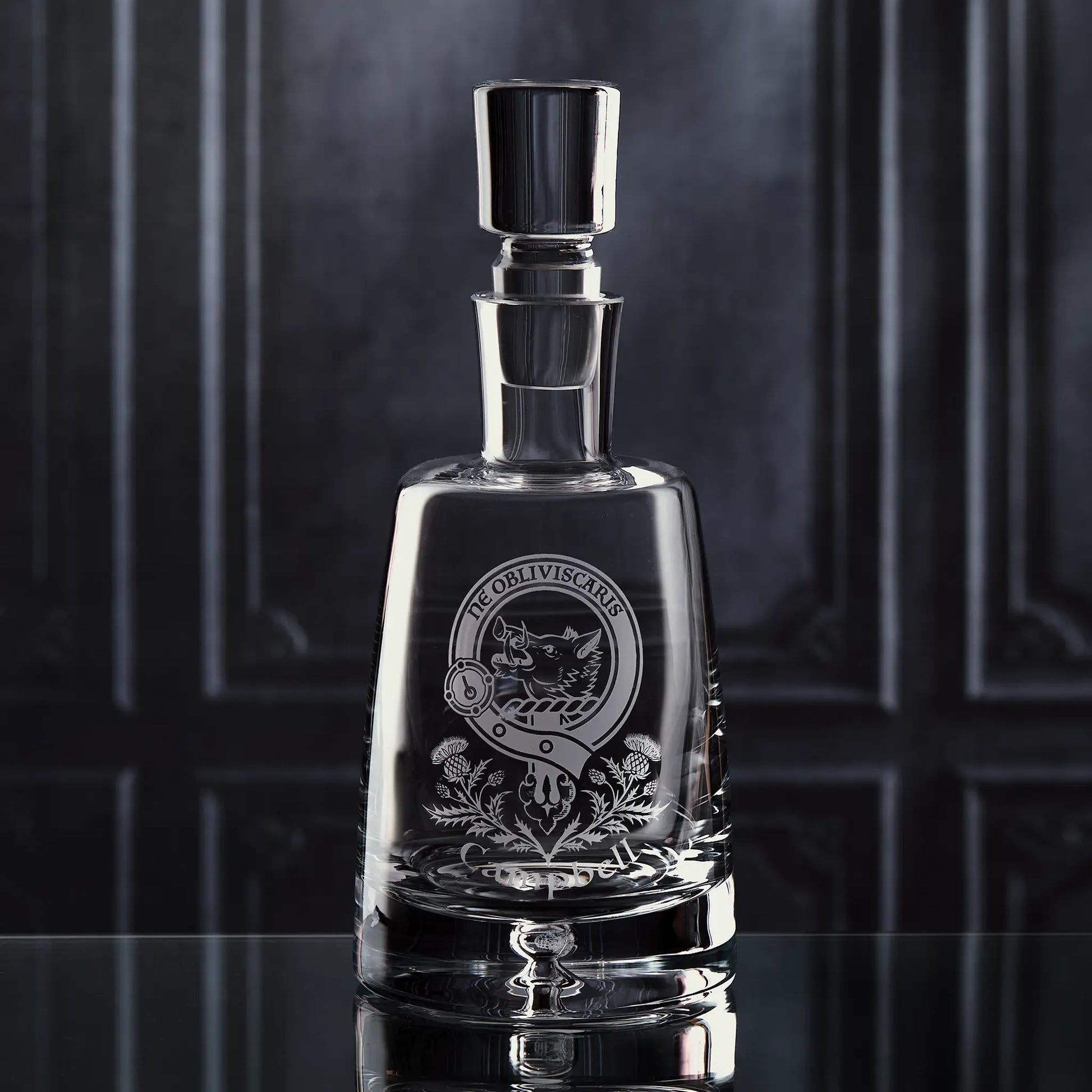 Handmade glass decanter engraved with personalised clan crest and thistle motif, shown on dark background to highlight shape and craftsmanship