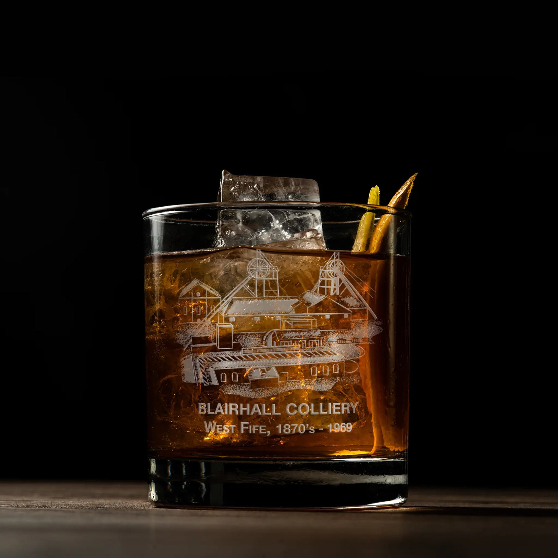 Glass of whiskey with ice featuring an engraved coal mine design on a dark background