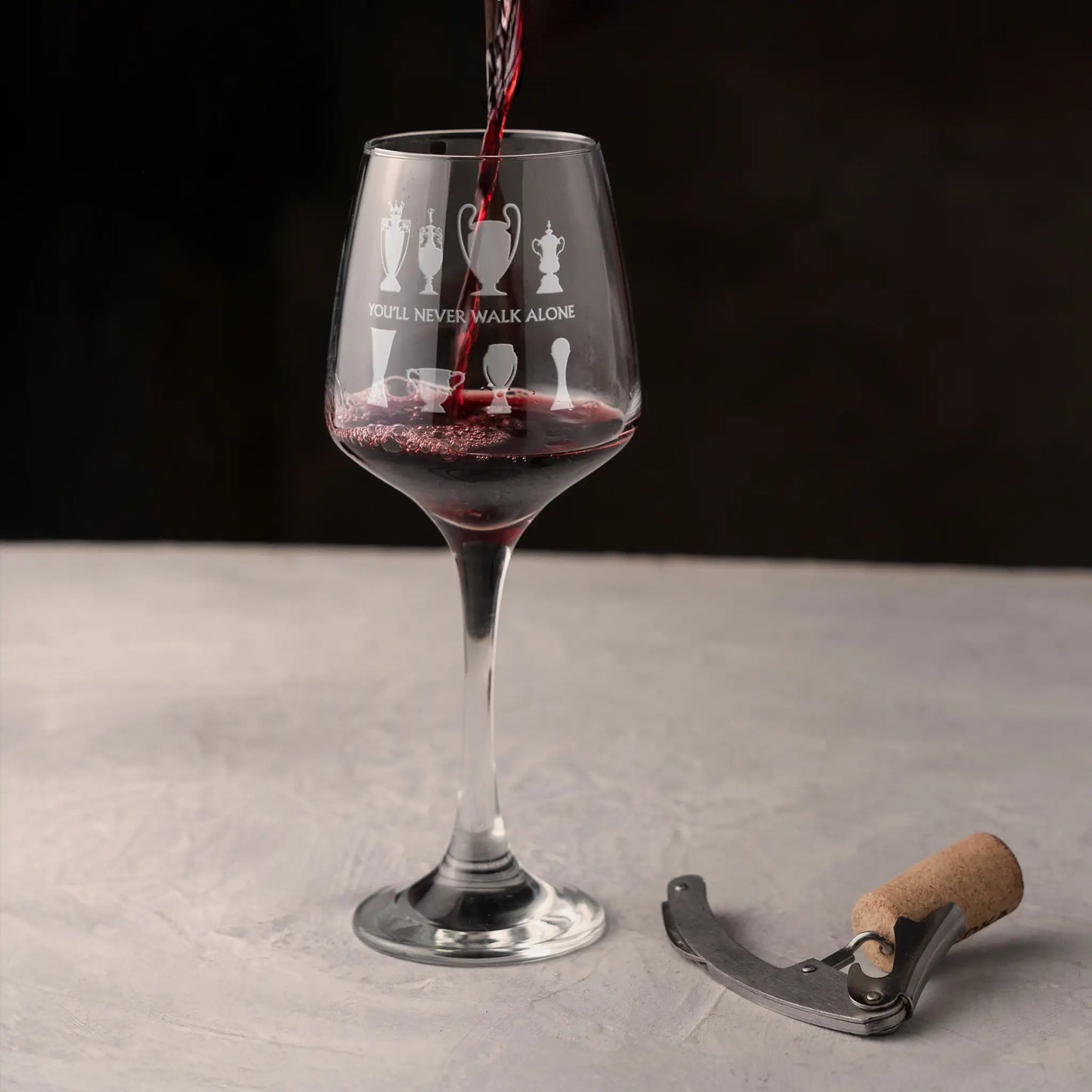 Wine glass with engraved football trophy design being filled with red wine on a grey surface against a dark background.