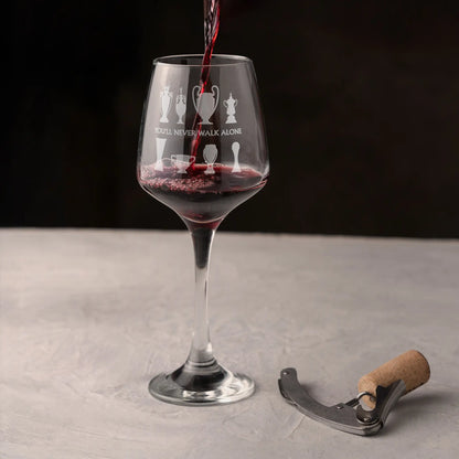 Wine glass with engraved football trophy design being filled with red wine on a grey surface against a dark background.