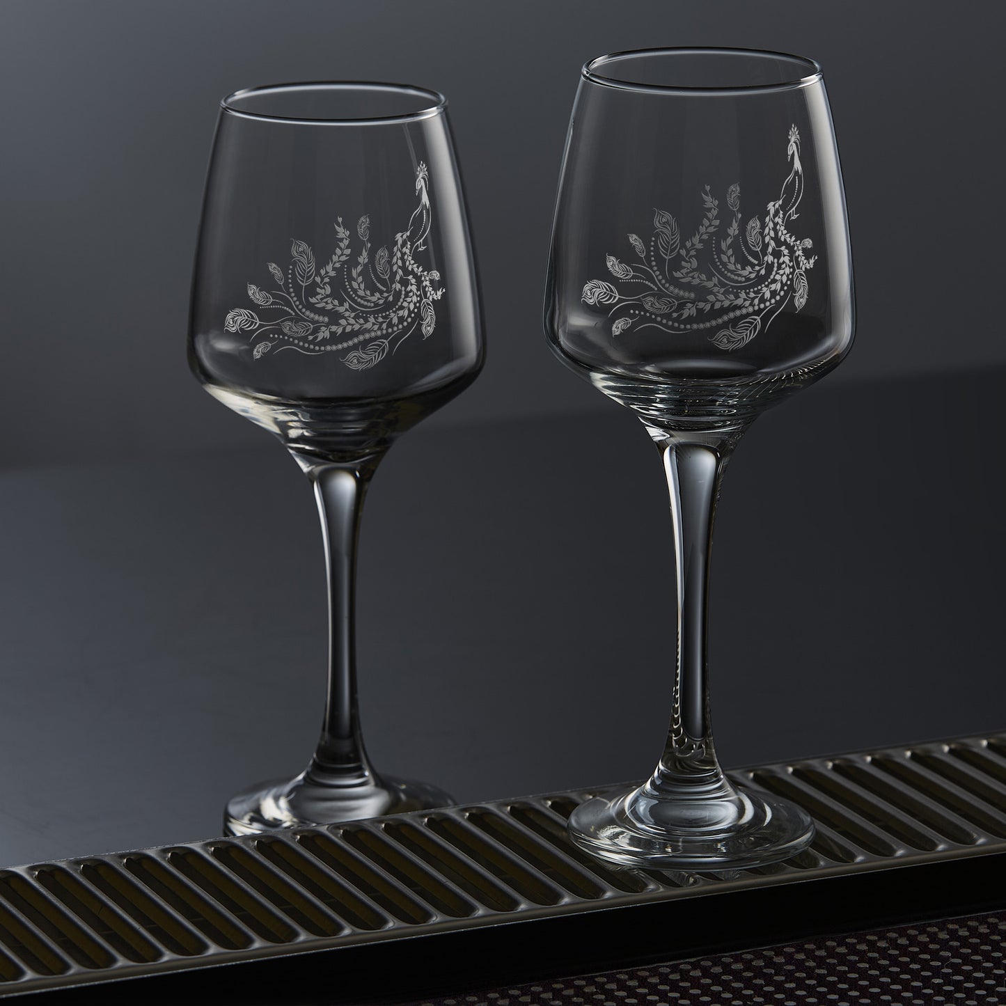Two wine glasses each featuring a single engraved peafowl with a flowing tail of feathers and trailing leaves, inspired by Pittencrieff Park’s peacocks