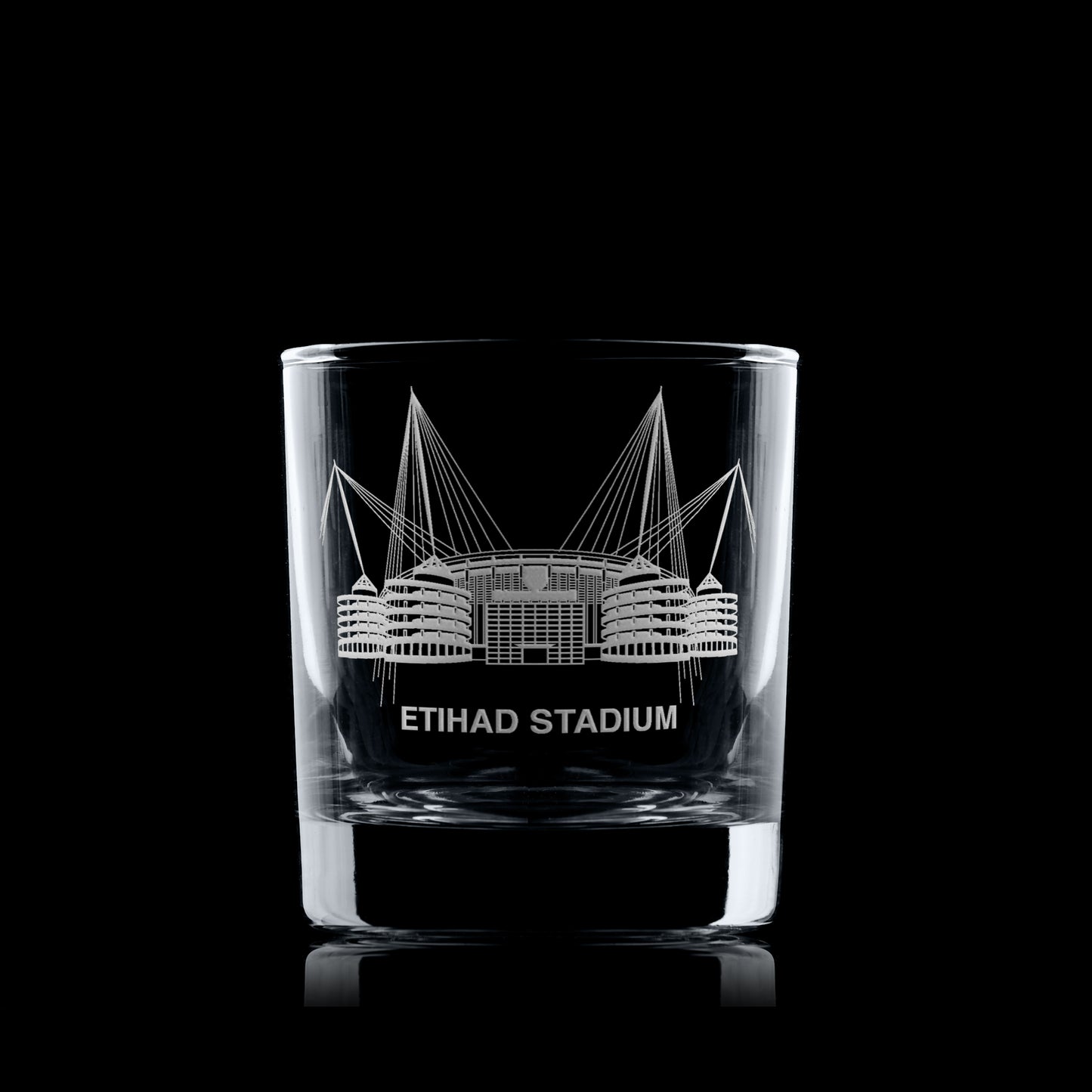 Whisky glass engraved with a line-art interpretation of the Etihad Stadium, photographed on a black background.