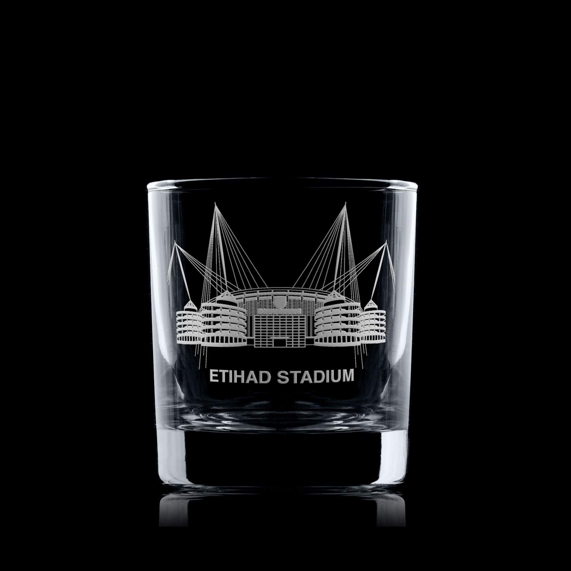 Whisky glass engraved with a line-art interpretation of the Etihad Stadium, photographed on a black background.
