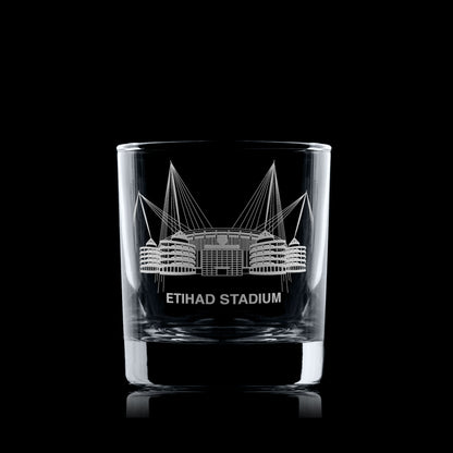 Whisky glass engraved with a line-art interpretation of the Etihad Stadium, photographed on a black background.
