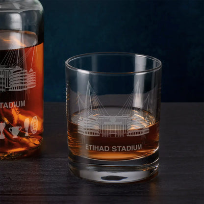Glass with etched Etihad Stadium design filled with amber liquid next to a bottle of the same liquid on a dark surface.