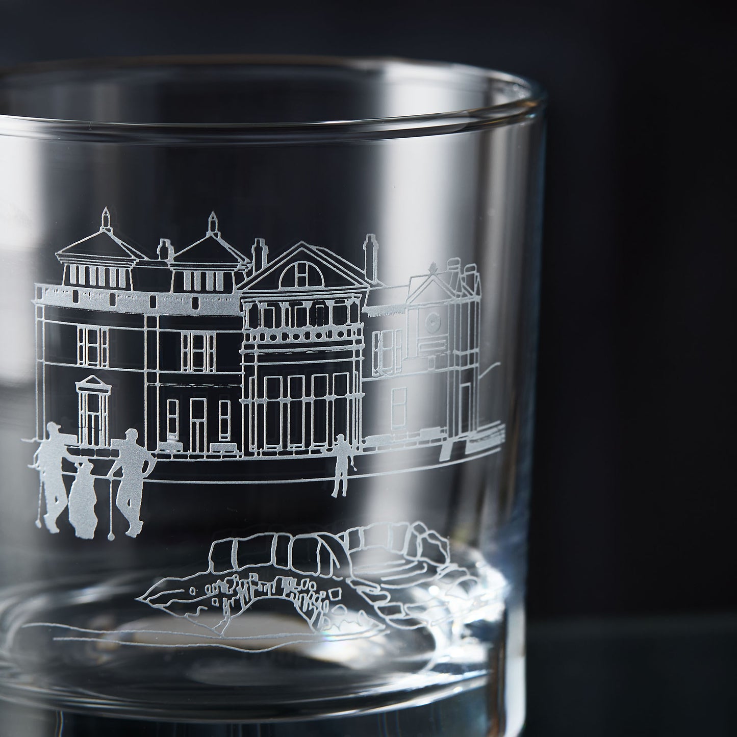 Glass whisky tumbler with engraved design of a golf clubhouse, bridge and three golfers on a dark background