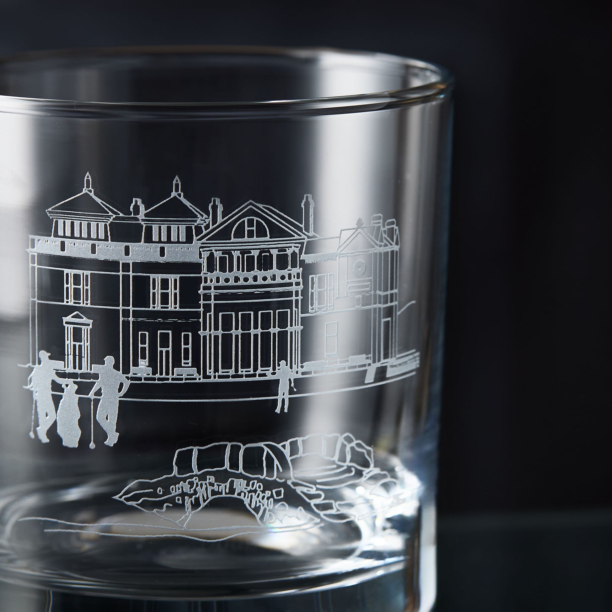 Glass whisky tumbler with engraved design of a golf clubhouse, bridge and three golfers on a dark background
