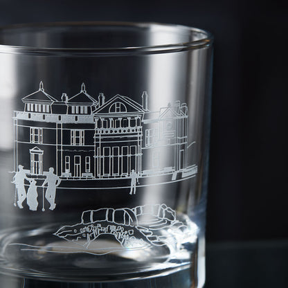 Glass whisky tumbler with engraved design of a golf clubhouse, bridge and three golfers on a dark background