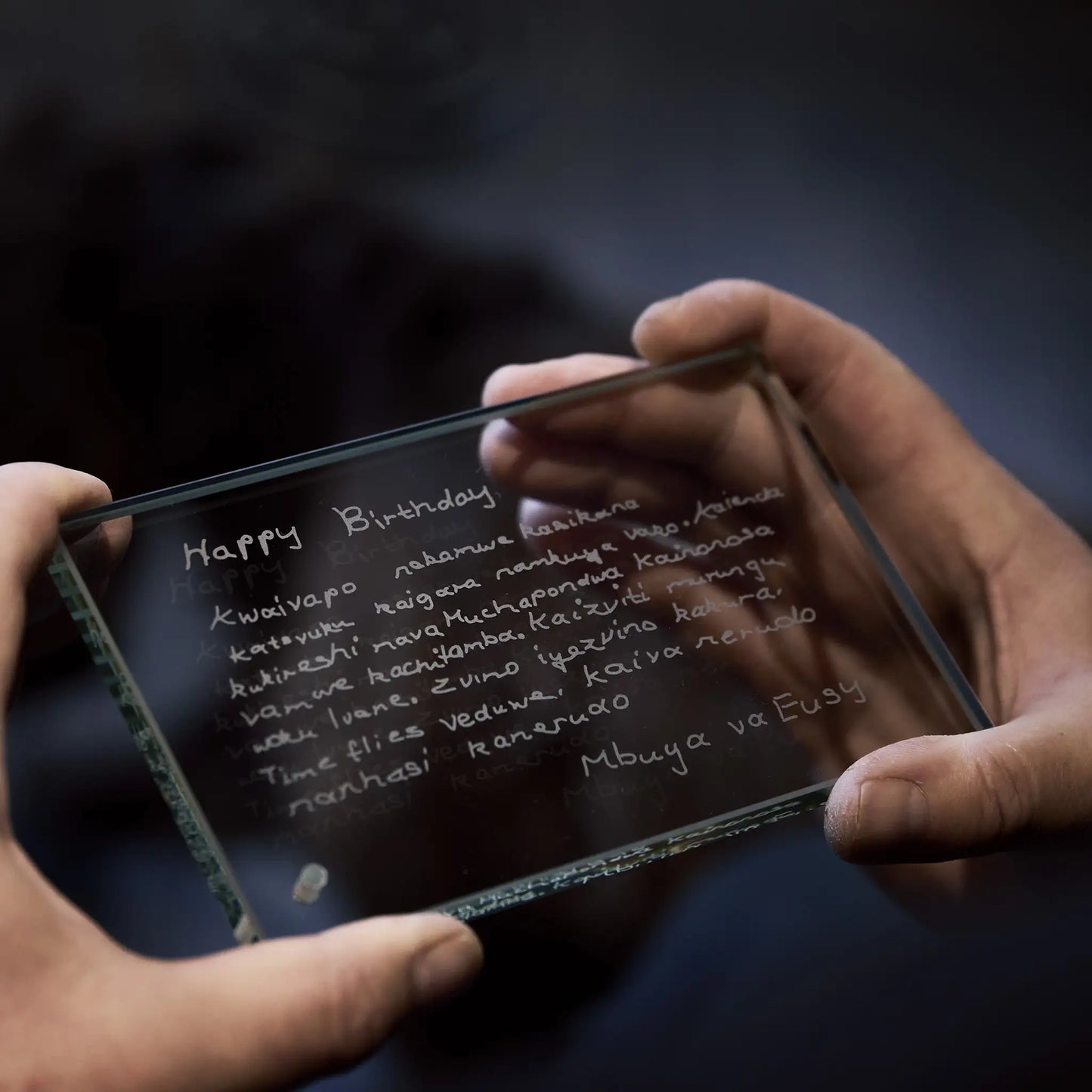 Hands holding a transparent glass with 'Happy Birthday' written on it against a dark background