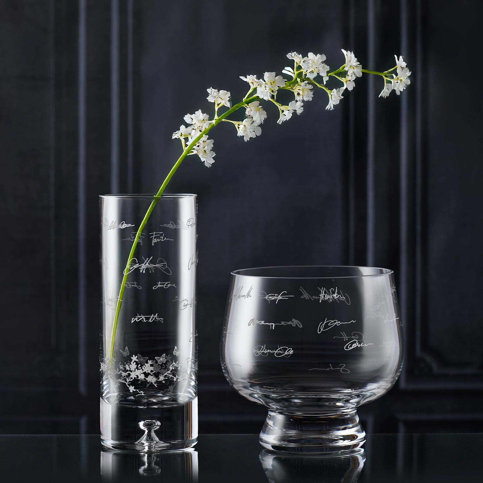 Two glass vases engraved with handwritten signatures against a dark background