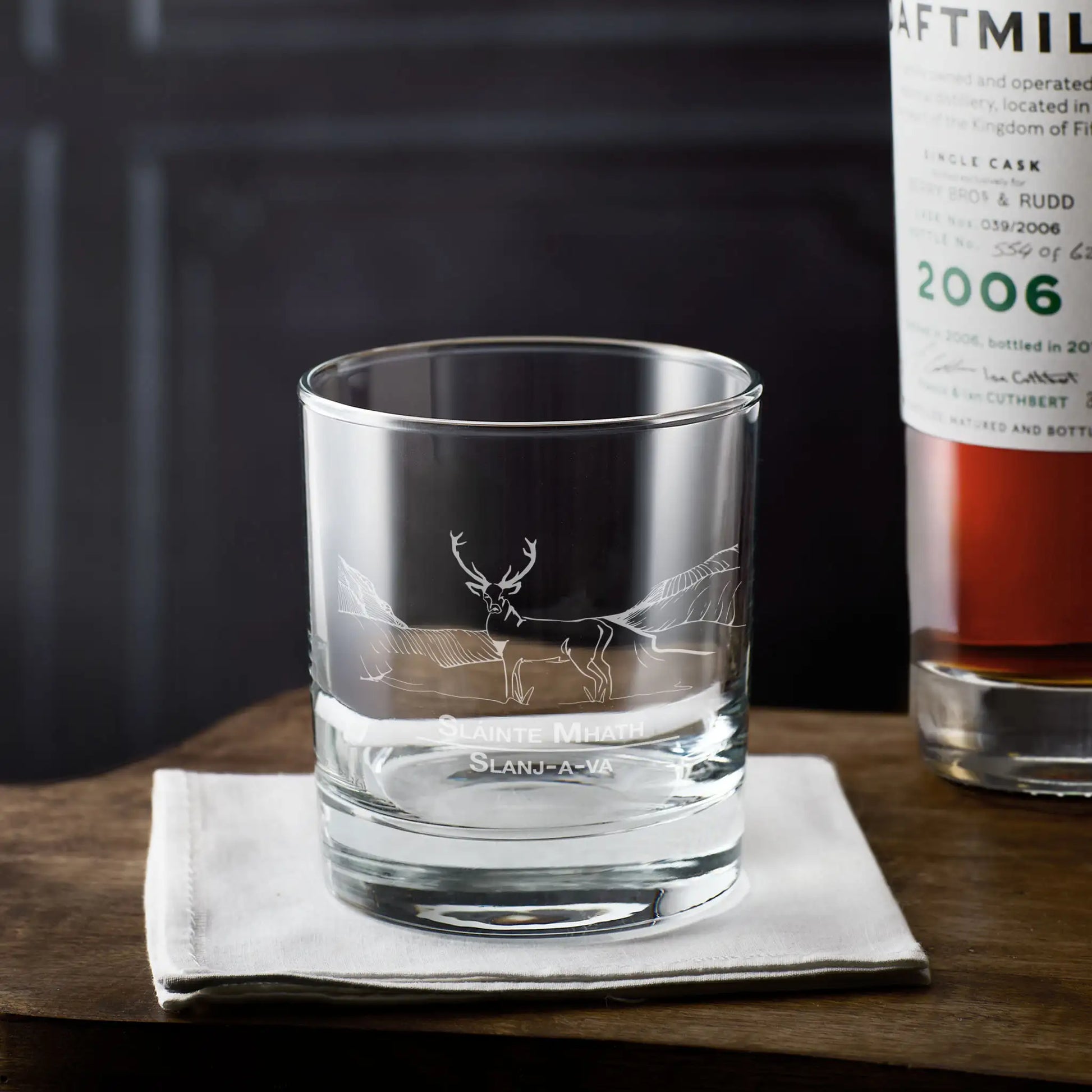 Whisky glass with engraved scottish stag and slainte design on a wooden surface next to a bottle of whisky.