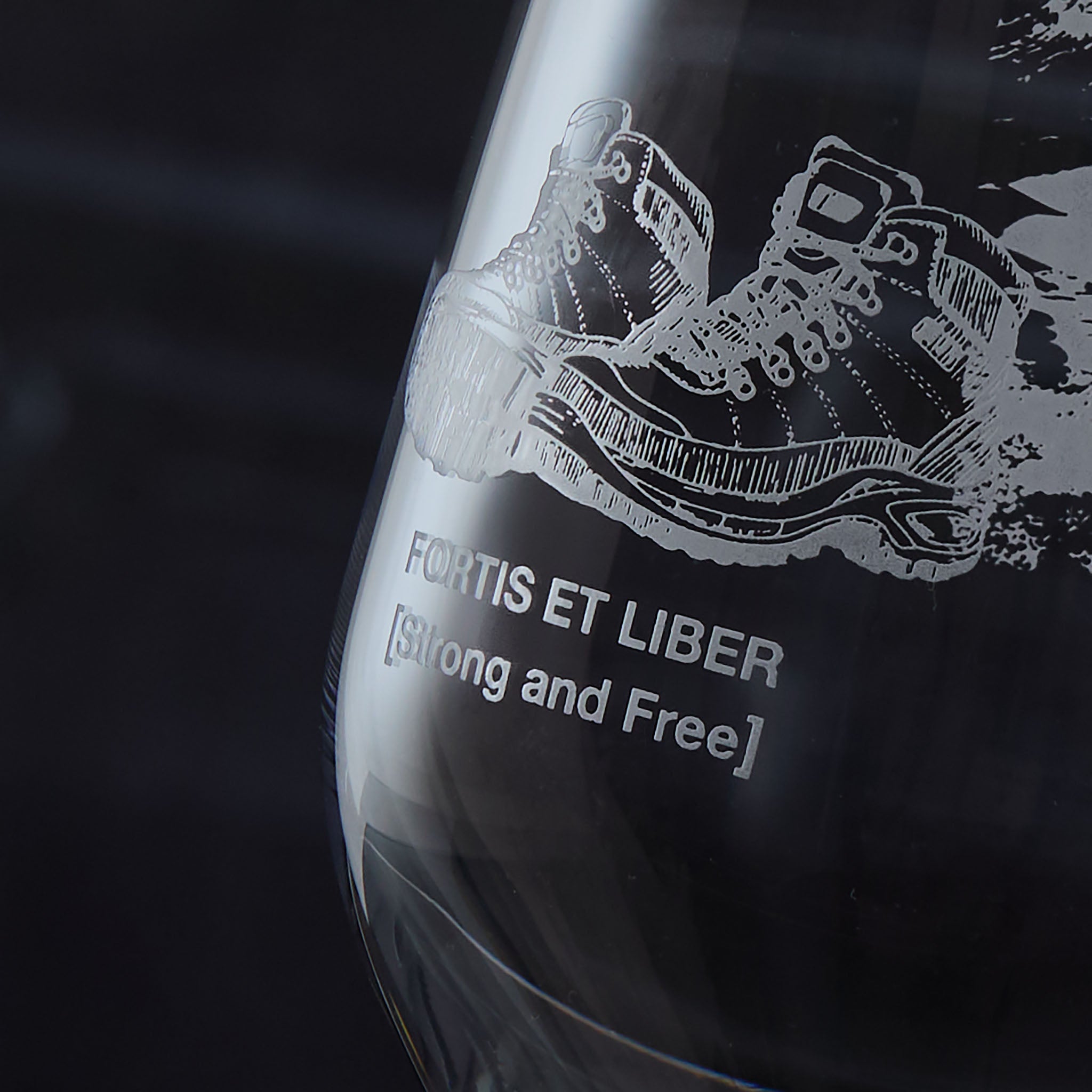 Mineral water glass engraved with walking boots and the text ‘Fortis Et Liber’ on a dark background”