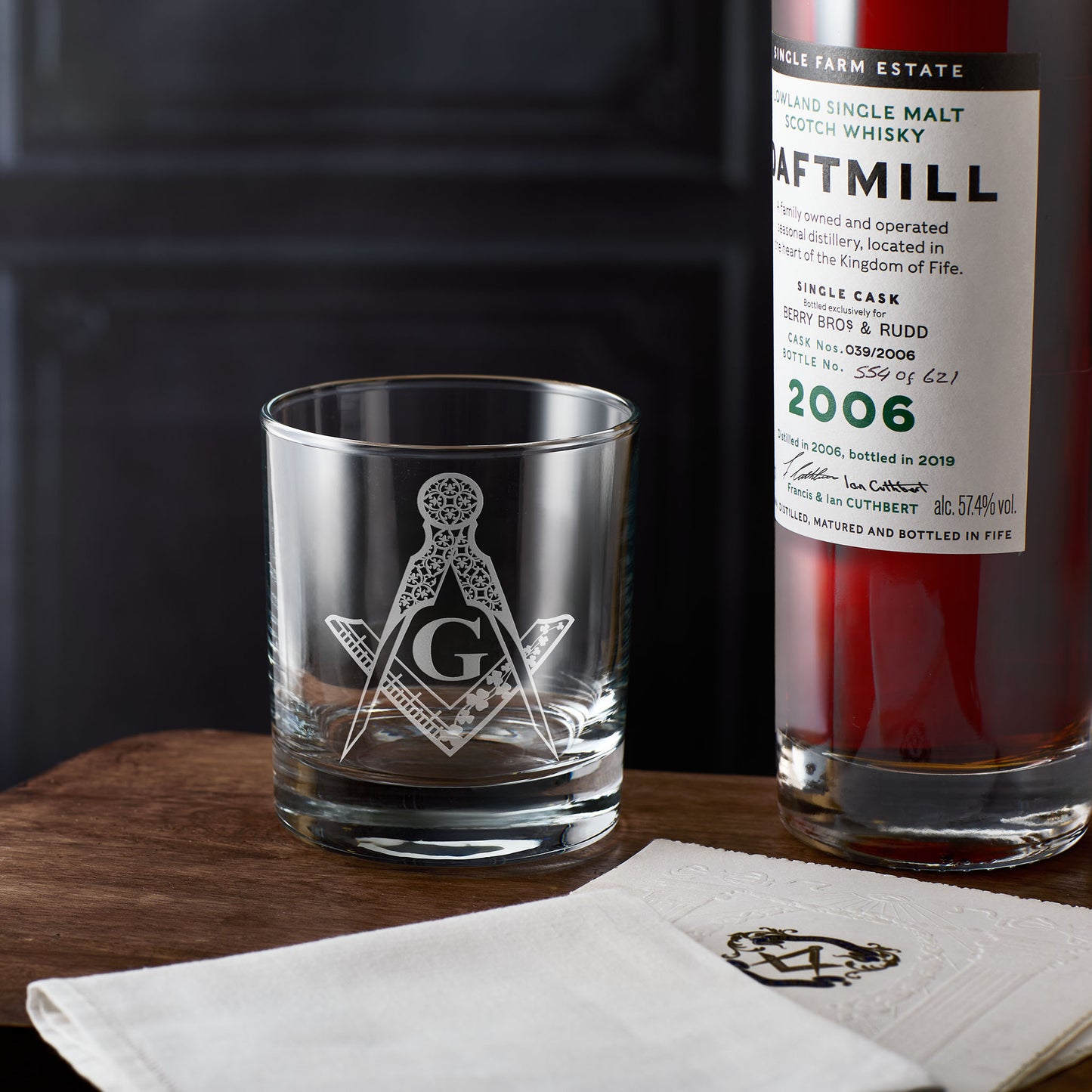 Whiskey glass with Masonic square and compass emblem next to a bottle of Daftmill whisky on a wooden surface.