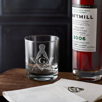Whiskey glass with Masonic square and compass emblem next to a bottle of Daftmill whisky on a wooden surface.