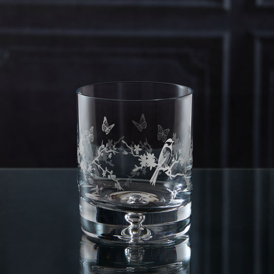 Clear glass tumbler with engraved garden birds, flowers and butterfly design on a dark surface.