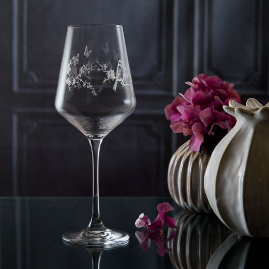 Wine glass with birds and a floral engraving on a reflective surface with flowers and a vase in the background.