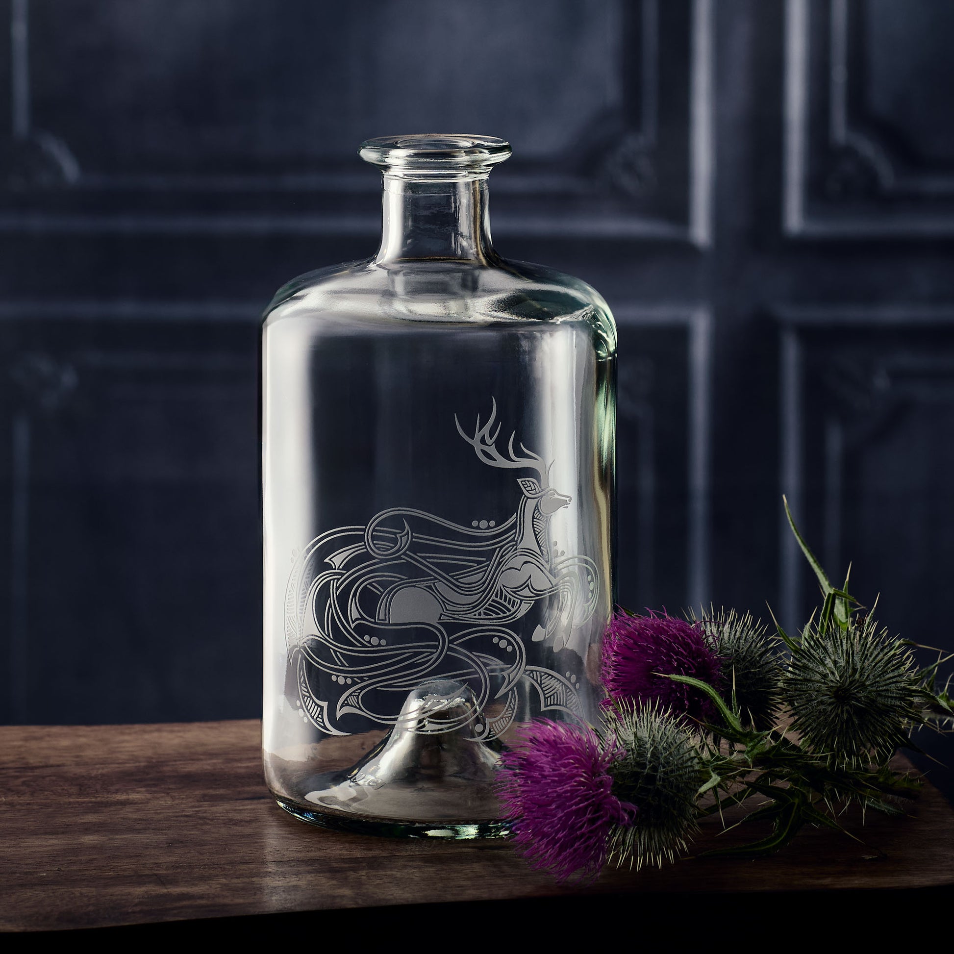 Whisky decanter with engraved celtic stag design on a wooden surface with purple flowers and greenery.