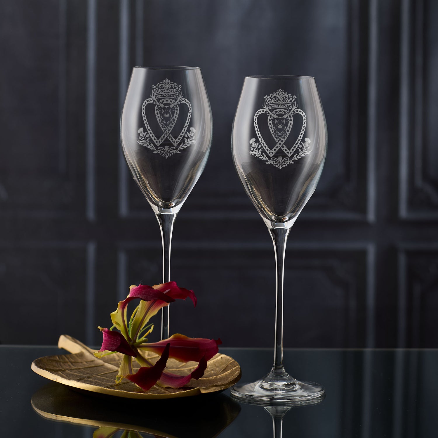 Two tall Atelier prosecco glasses engraved with crowned Luckenbooth, thistles, and stag design, shown on dark background to highlight engraving detail