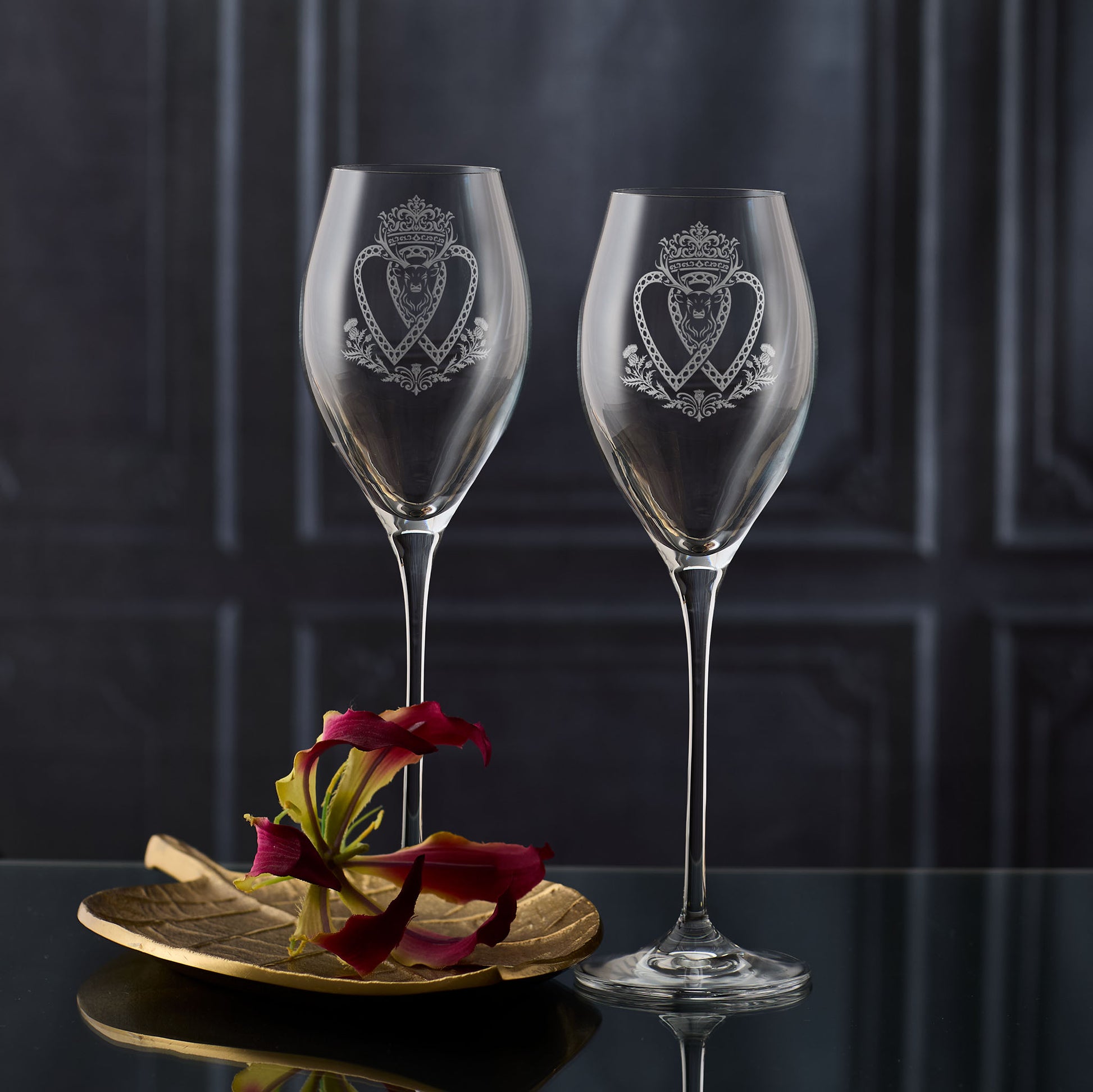 Two tall Atelier prosecco glasses engraved with crowned Luckenbooth, thistles, and stag design, shown on dark background to highlight engraving detail