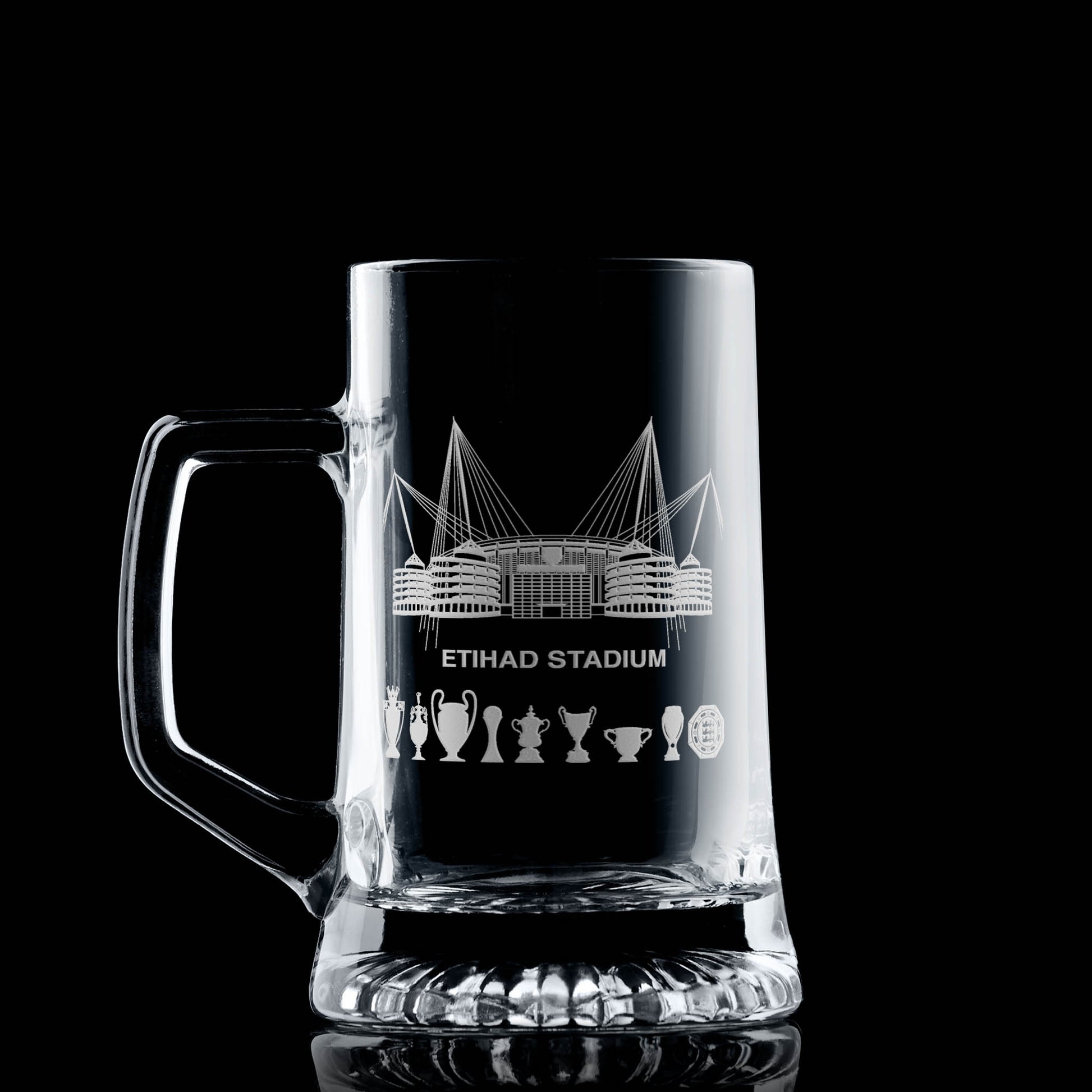 Clear glass tankard engraved with a line-art interpretation of the Etihad Stadium and trophy silhouettes, photographed on a dark background.