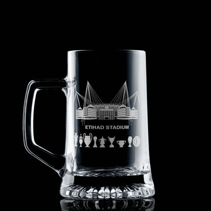 Clear glass tankard engraved with a line-art interpretation of the Etihad Stadium and trophy silhouettes, photographed on a dark background.
