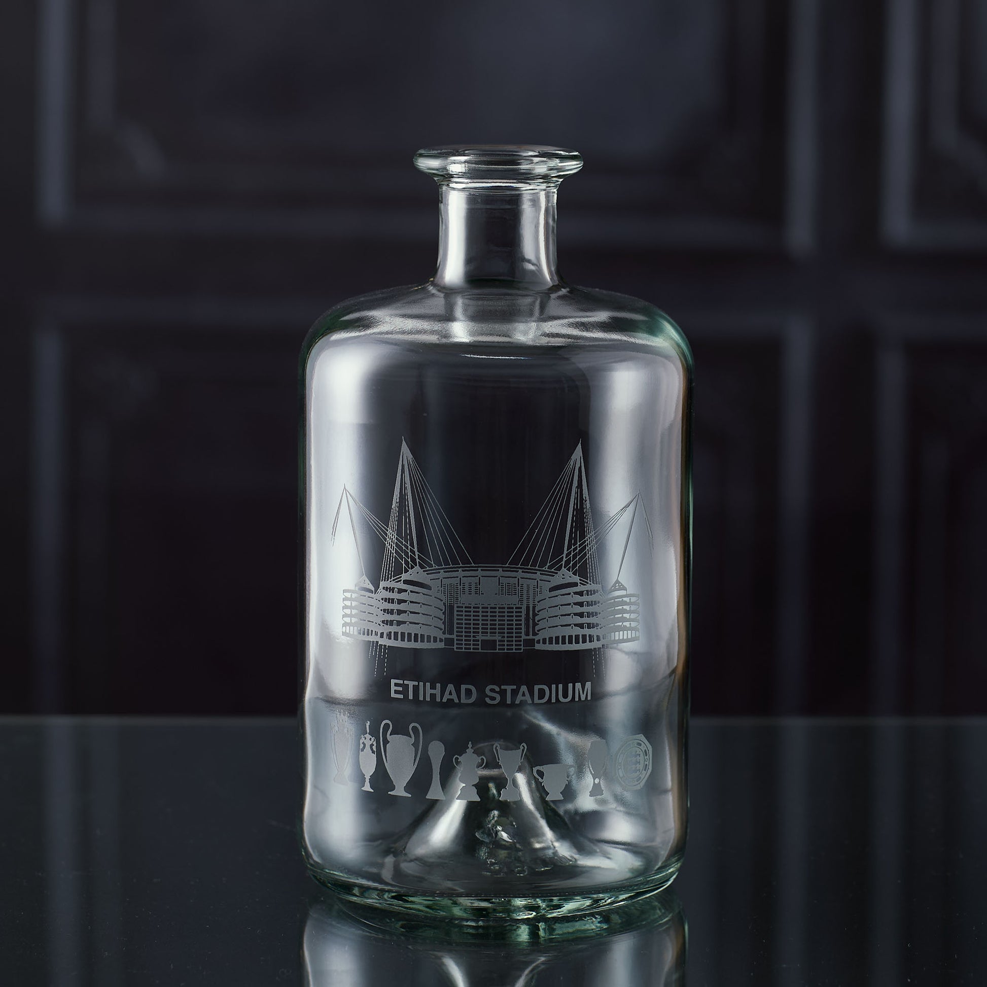 Apothecary-style glass decanter engraved with a line-art interpretation of the Etihad Stadium and trophy silhouettes, photographed on a dark background.