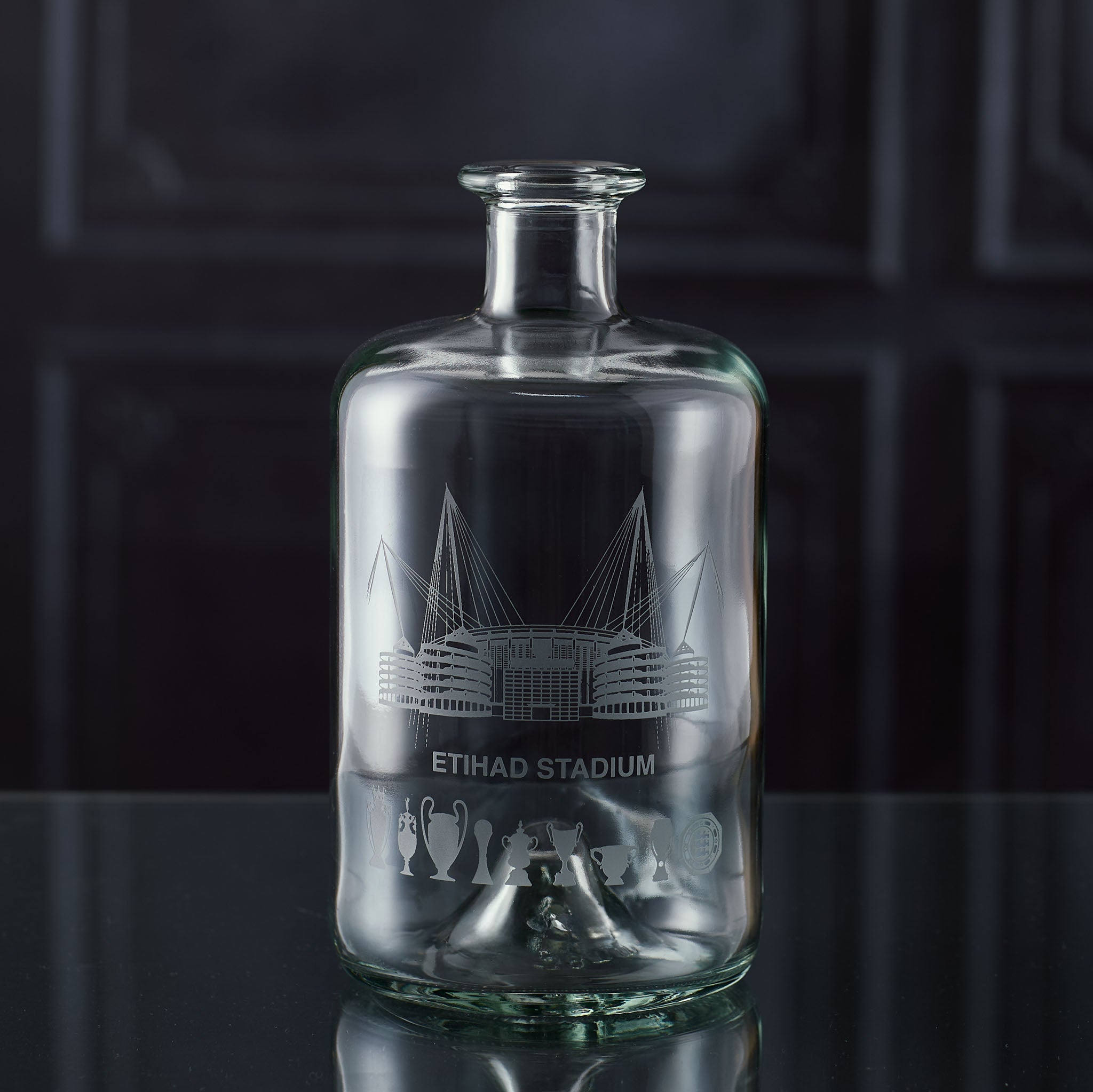 Apothecary-style glass decanter engraved with a line-art interpretation of the Etihad Stadium and trophy silhouettes, photographed on a dark background.